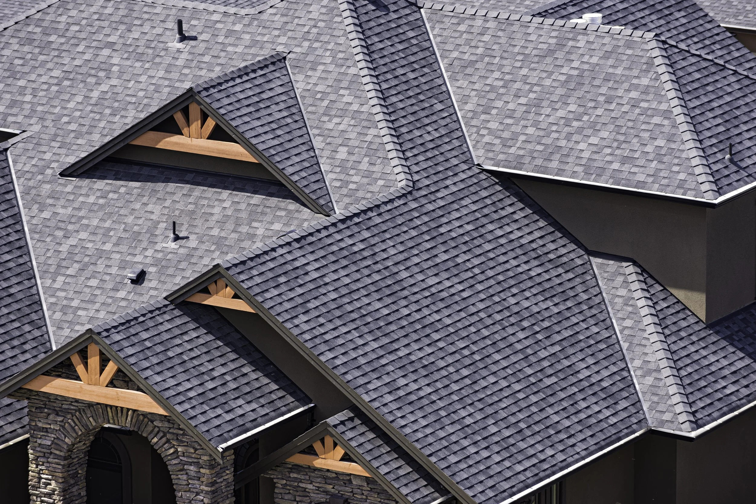 Close-up aerial view of a house with gray shingle roof, dormer windows, stone facade, and wooden trim