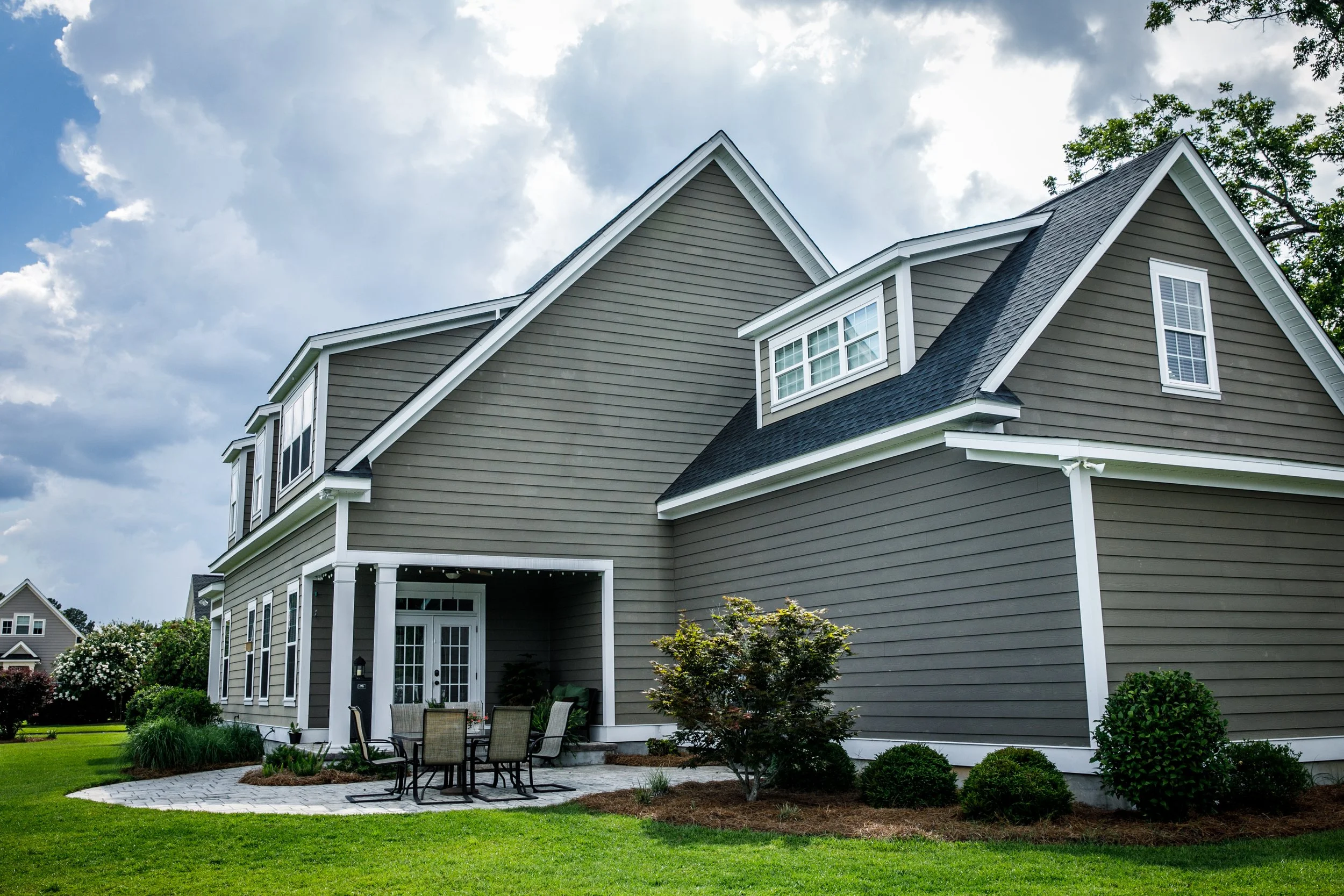 A two-story house with gray siding, multiple roof peaks, white trim, and a covered back patio with outdoor furniture, surrounded by a well-maintained lawn and landscaped garden under a cloudy sky.