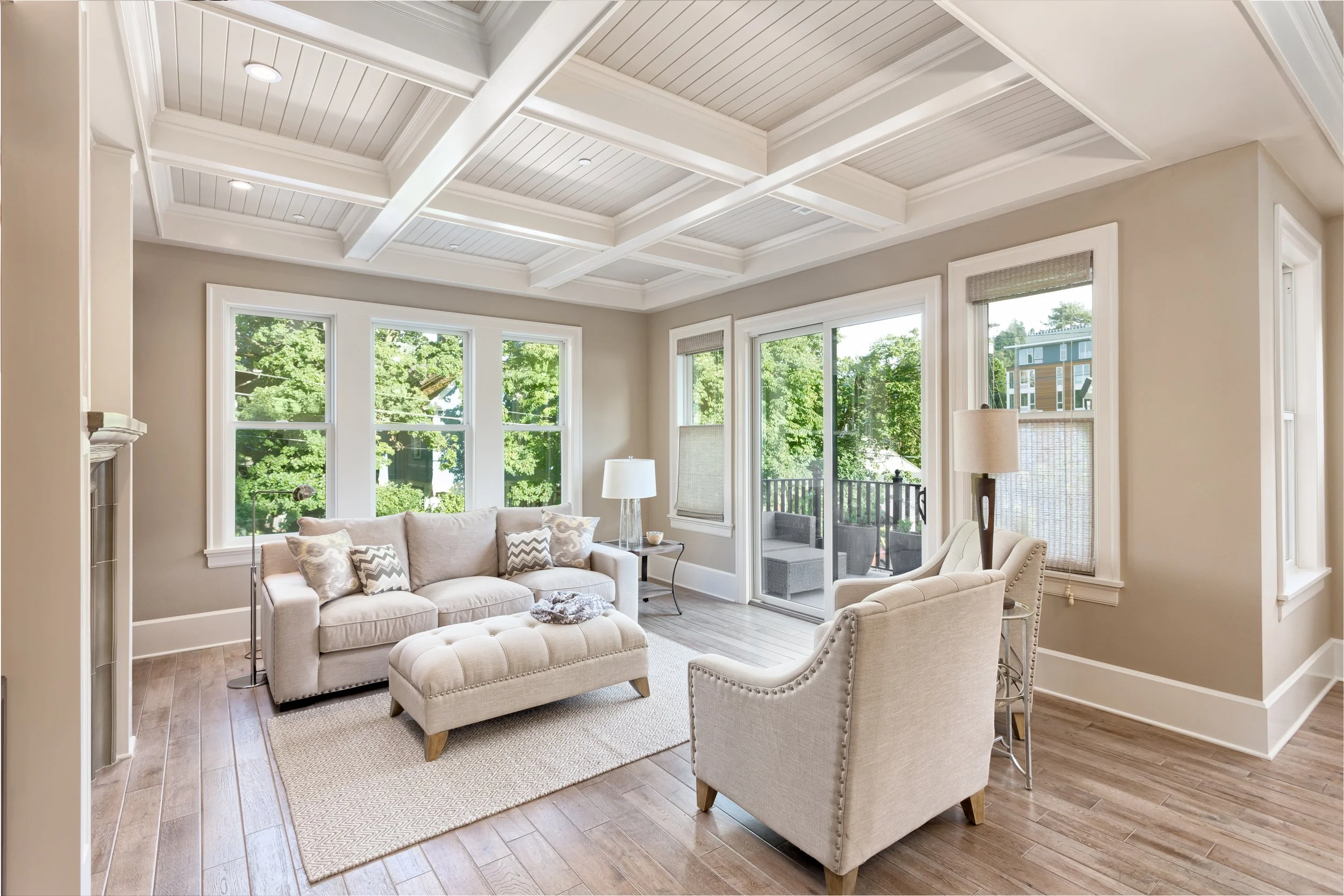 Living room with large windows, beige sofa with cushions, white armchair, side table with a lamp, light wood flooring, coffered ceiling, and patio door leading to balcony.