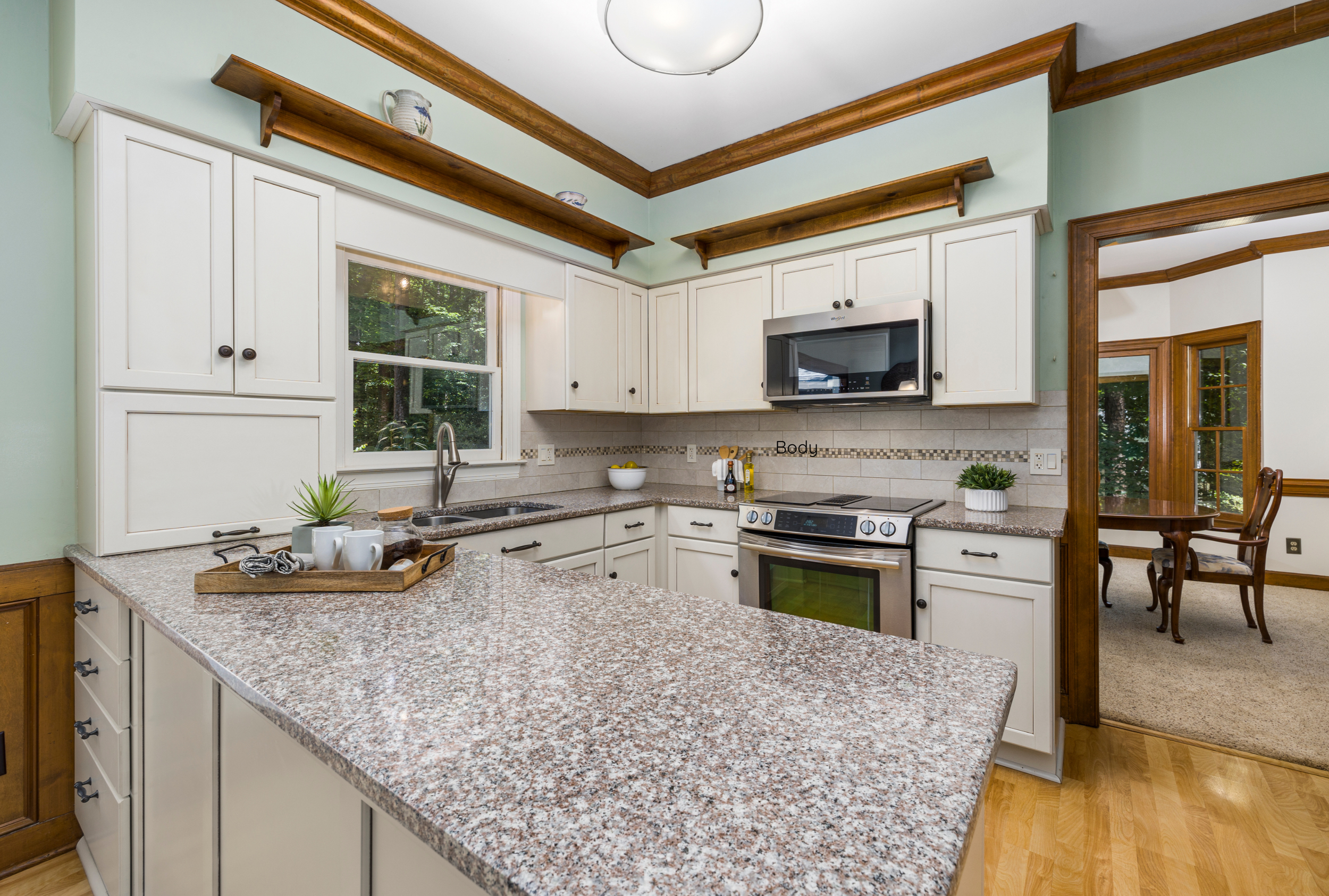 A spacious kitchen with frosted cabinets, a granite countertop island, a stainless steel microwave, a window with a view of trees, and wooden accents including crown molding and shelves.