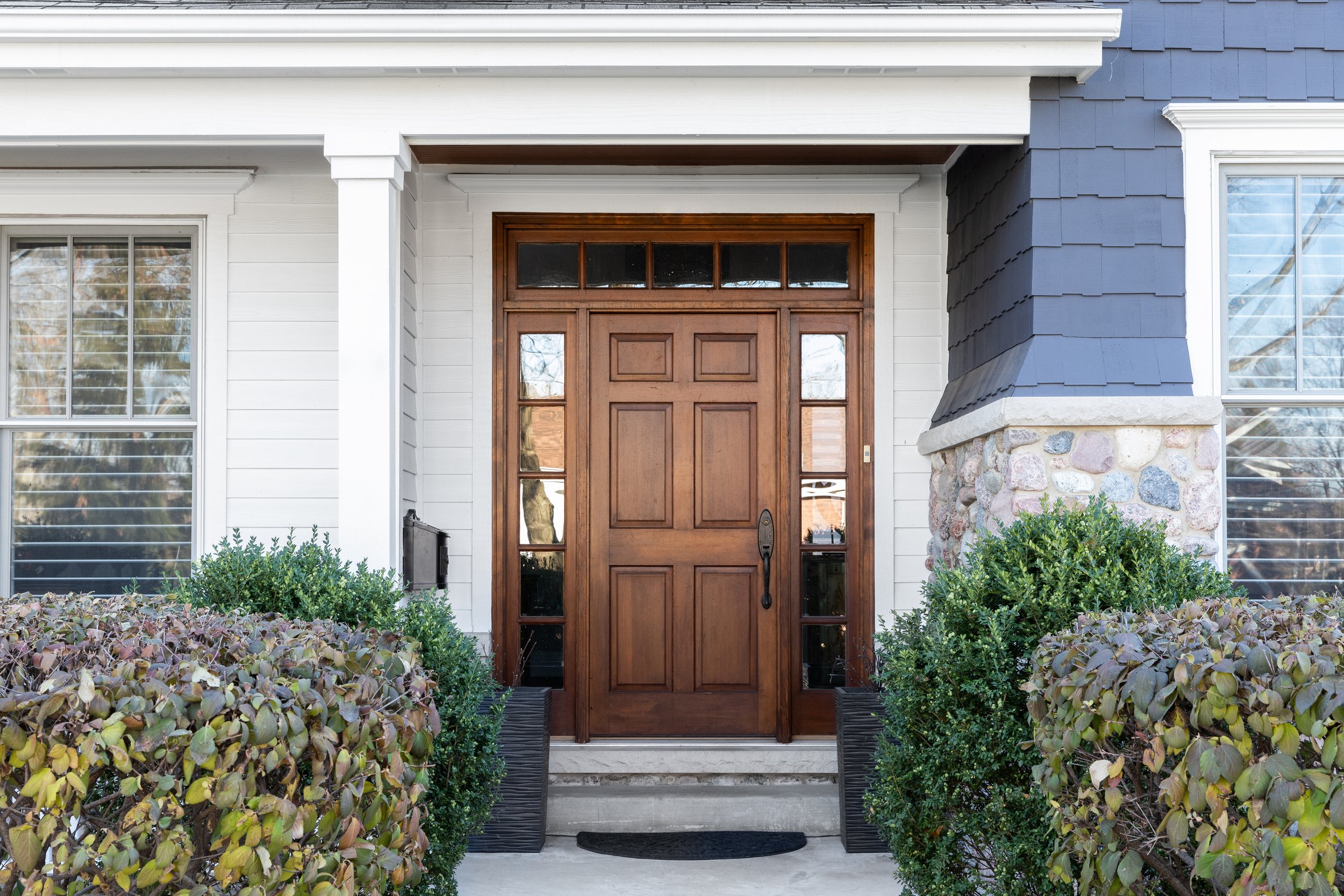 Front door of a house with a wooden door, tall glass side panels, and a small window above, surrounded by bushes and a stone foundation.