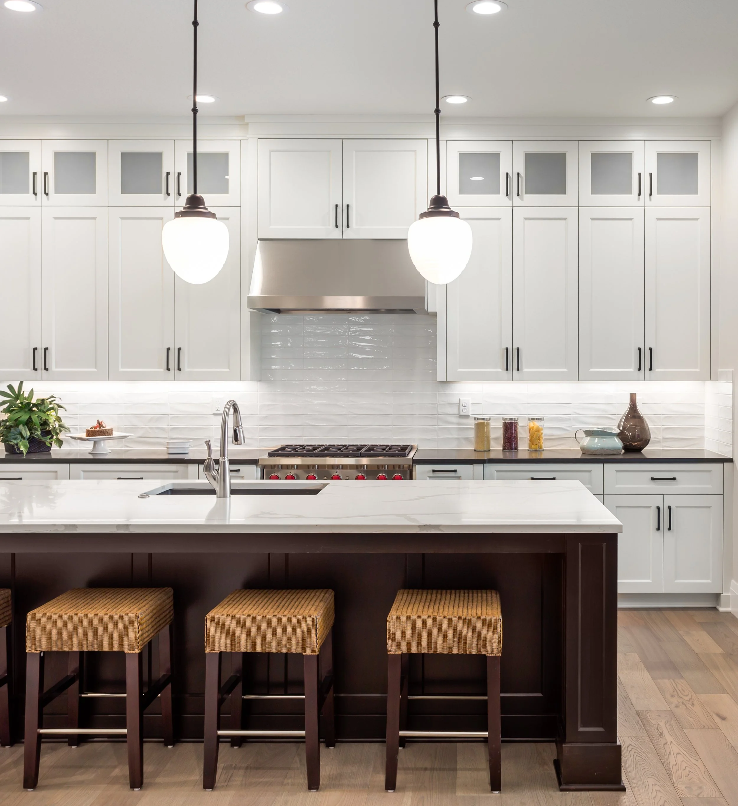A modern kitchen featuring white cabinetry, a white island with a built-in sink, dark wood base, and four wicker stools underneath. The backsplash is white subway tiles, and there are decorative items on the counter such as a small cake, jars, and a ceramic vase. Overhead, there are two pendant lights with white glass shades.
