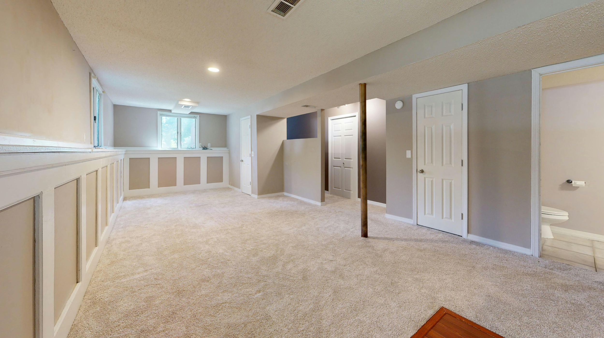 A spacious, empty basement room with beige carpet, white and beige paneled walls, and multiple windows letting in natural light. There are several doors, a support pole, and a small section of bathroom visible.