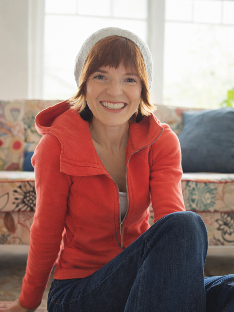 A woman with short red hair wearing a white knit hat, red hoodie, and blue jeans sitting on the floor in a brightly lit living room, smiling at the camera.