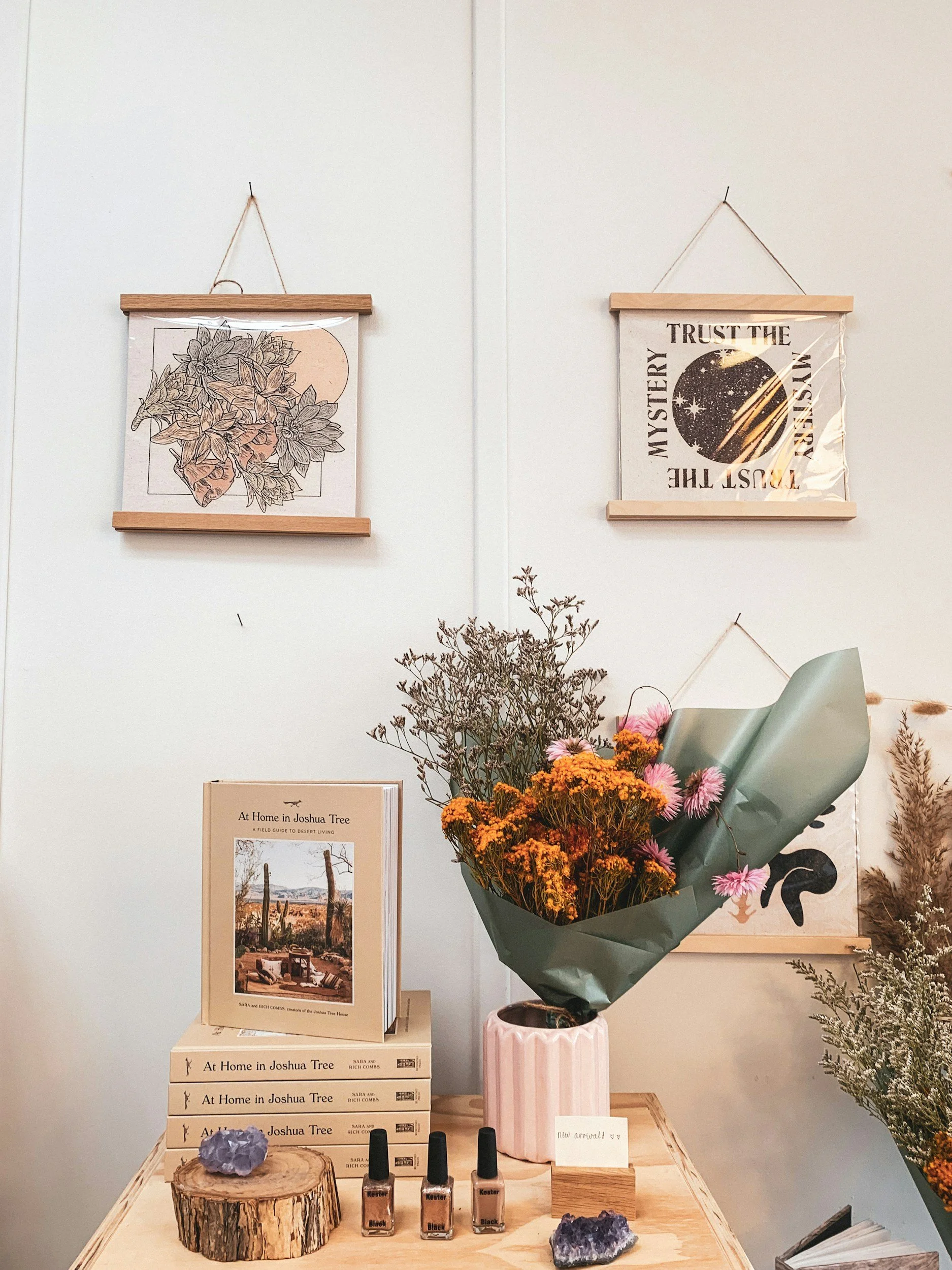 Display of books, a bouquet of flowers in a pink vase, decorative crystals, and nail polish bottles on a wooden table, with framed artwork and informational posters on the wall.