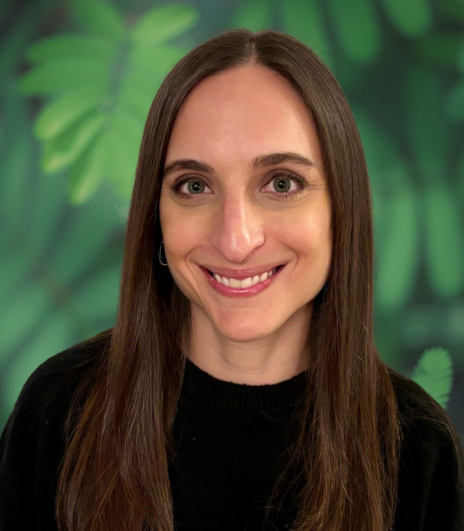 A woman with long brown hair, smiling, wearing a black top, against a green leafy background.