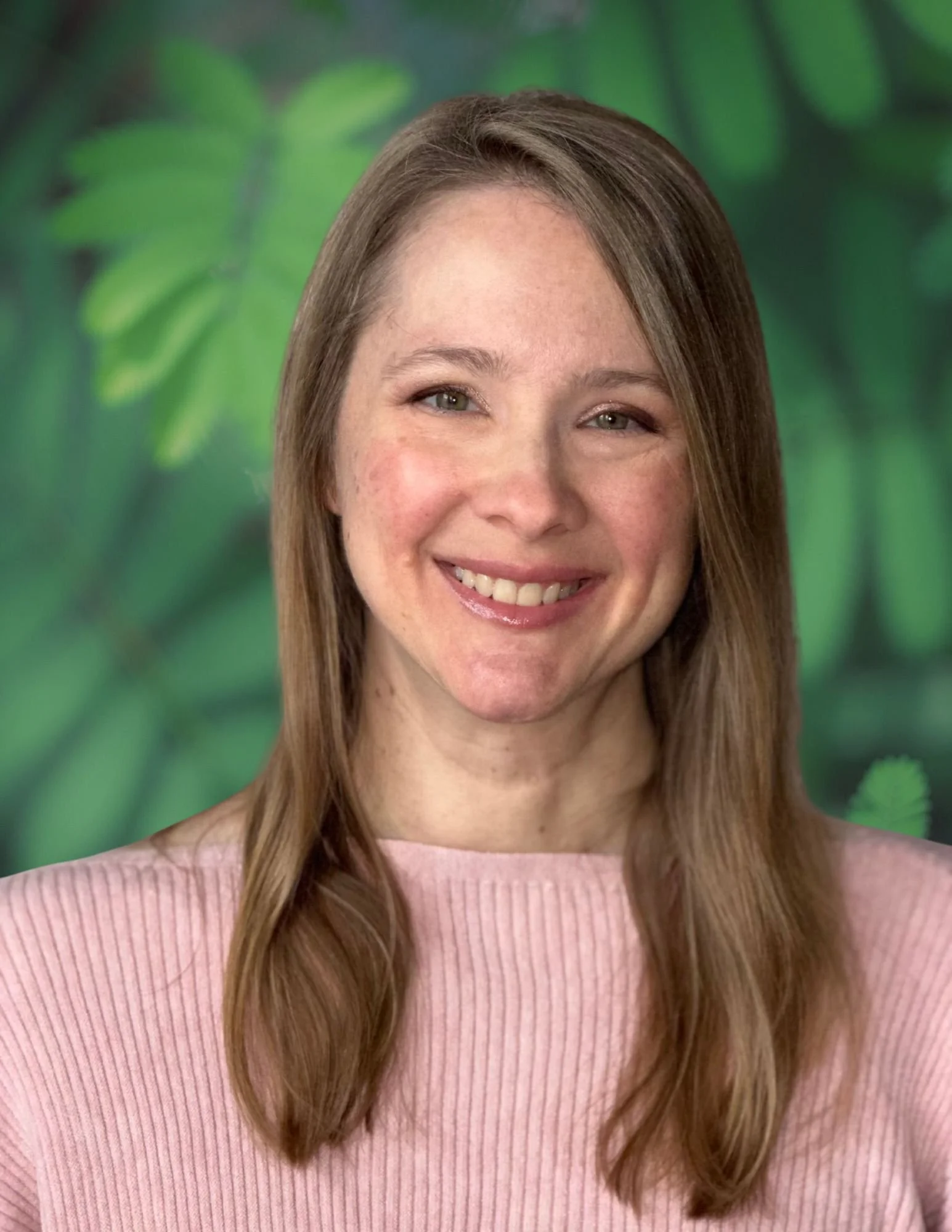 A woman with long light brown hair and light skin, smiling, wearing a pink ribbed sweater, with a green leafy background.