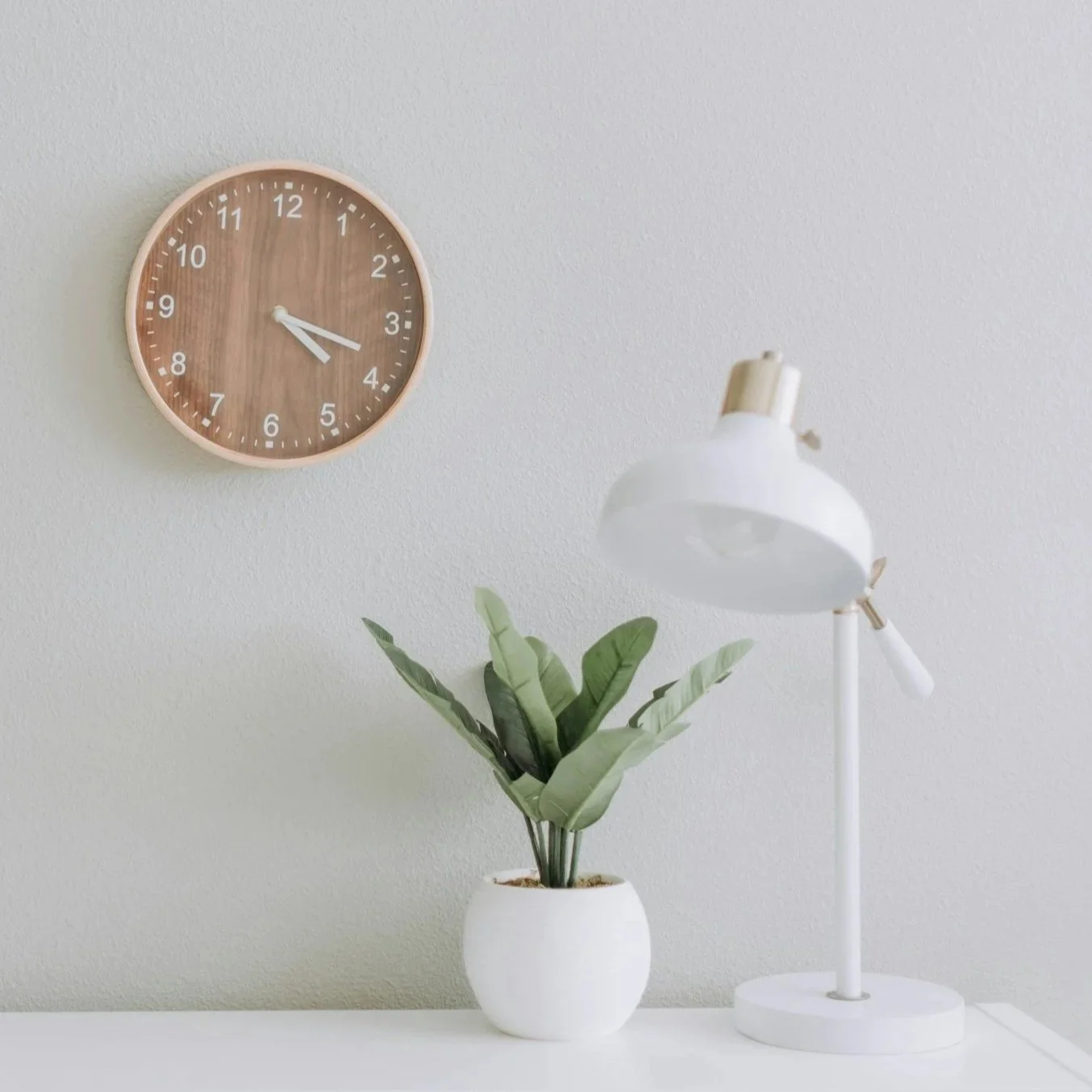 A wall clock with a wooden face and white numbers, a potted plant with green leaves, and a white desk lamp on a white surface against a plain light-colored wall.