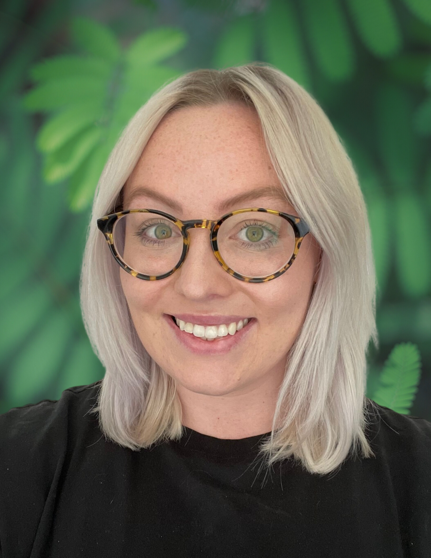 A woman with blonde hair, green eyes, wearing tortoiseshell glasses, smiling, against a green leafy background.