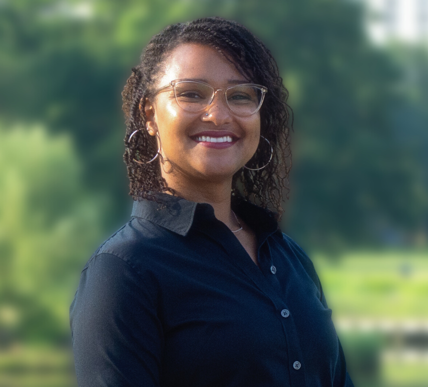 Smiling woman with glasses and hoop earrings against a green leafy background.