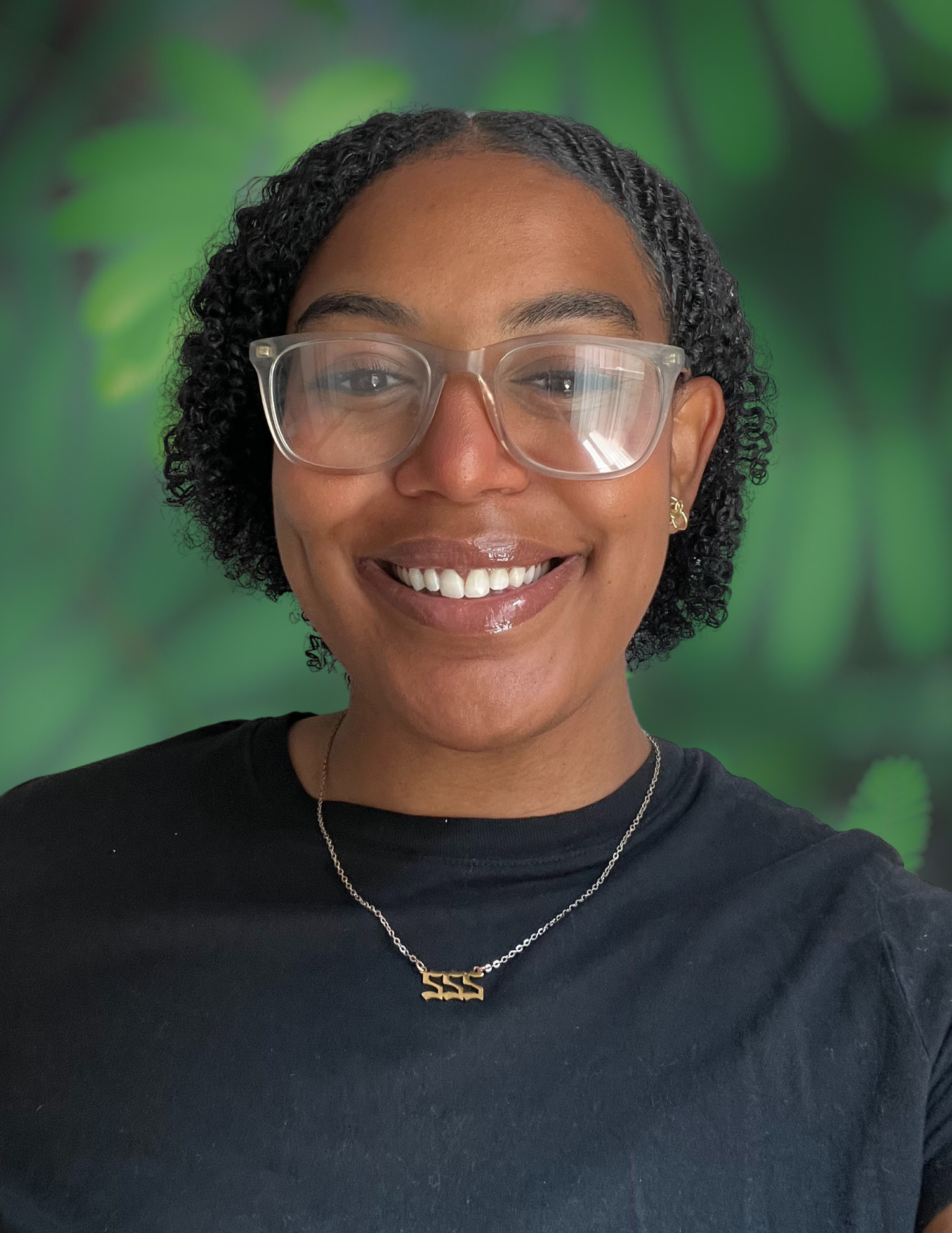 A woman with curly black hair, glasses, smiling, wearing a black shirt, and a gold necklace against a green leafy background.