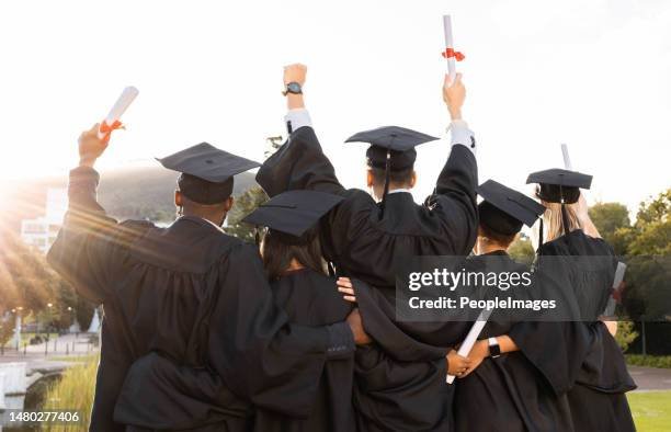 Group of graduates in caps and gowns celebrating at graduation ceremony, holding diplomas in the air.