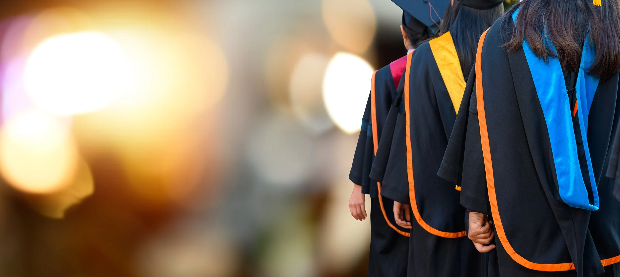 A line of graduates wearing black gowns with orange and blue accents, standing with their hands at their sides, with a blurred background featuring bright lights.