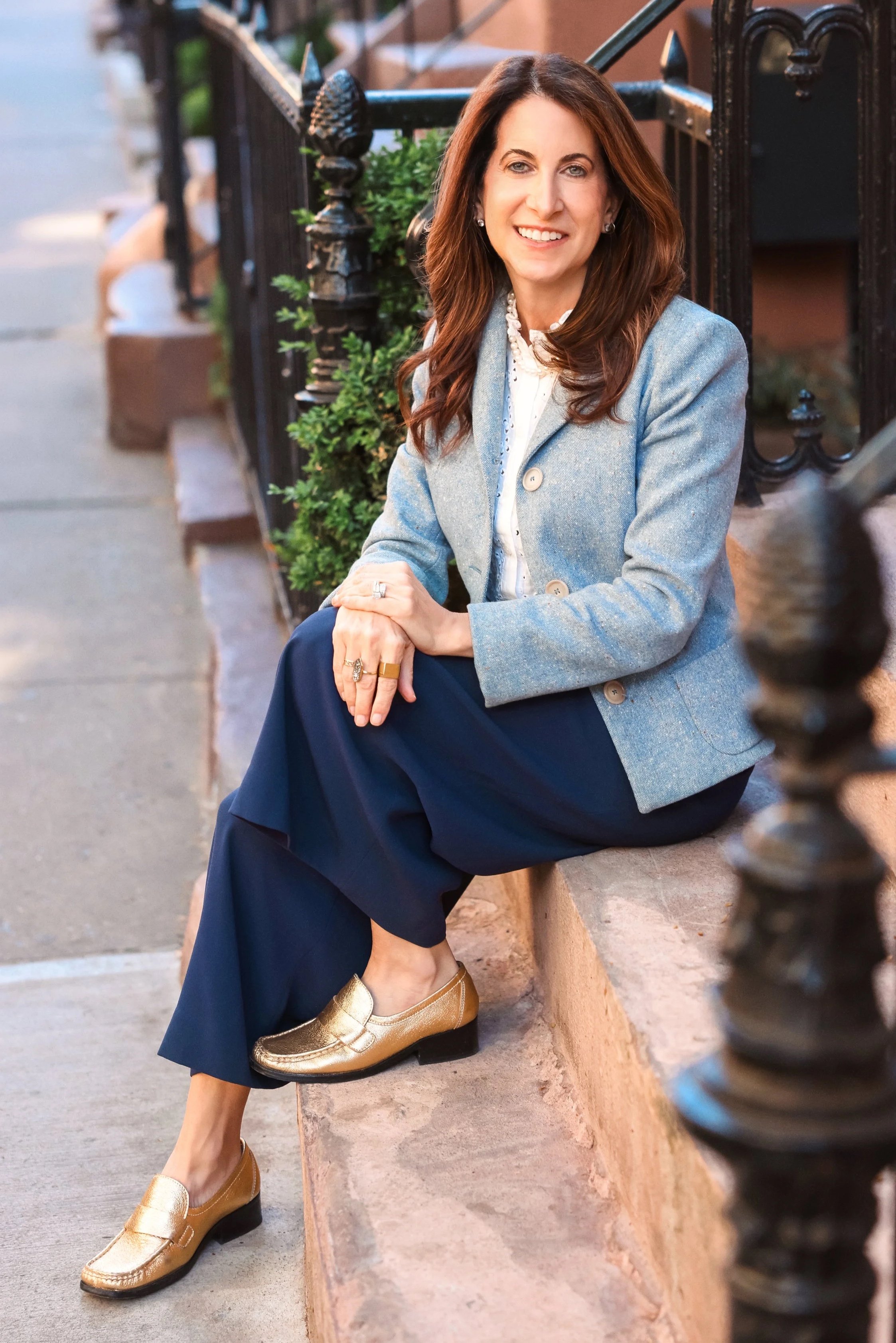 A woman with long brown hair and light skin is smiling while sitting on a concrete ledge on a city sidewalk. She is wearing a light blue blazer, a white blouse, navy blue pants, and gold loafers. She has jewelry including rings, earrings, and a necklace. There are greenery and a black iron fence behind her, and the background shows a townhouse or apartment building.