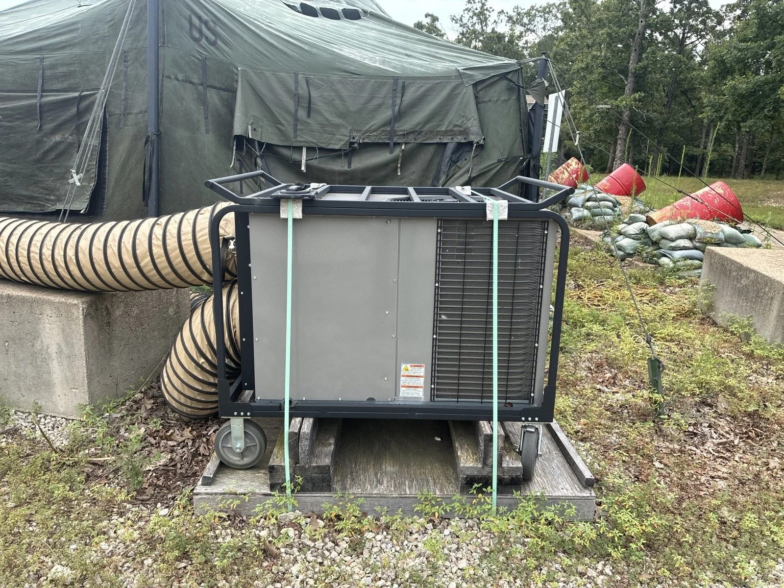 Portable air conditioning unit outdoors on a wooden platform next to a large green military-style tent with trees in the background.