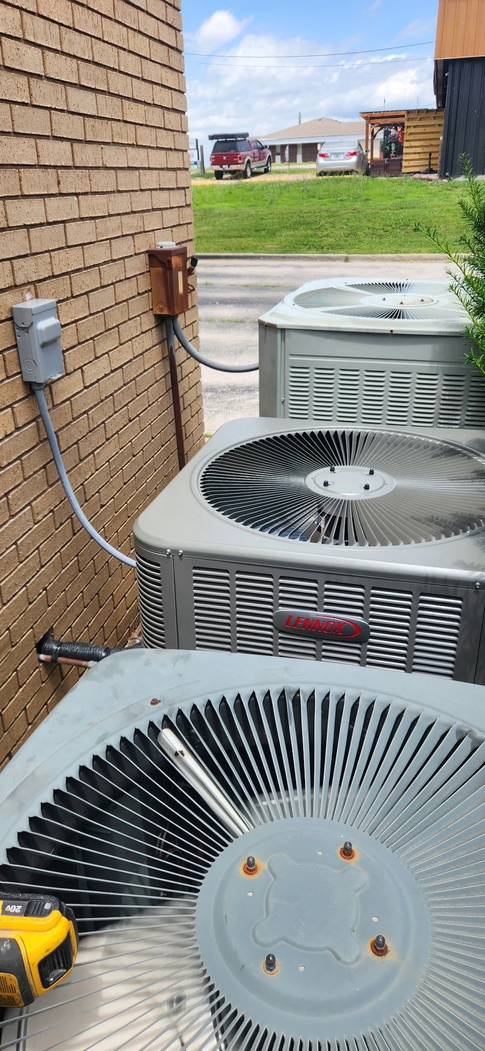 Three outdoor air conditioning units installed against a brick wall outside a building, with one unit disassembled and tools nearby.