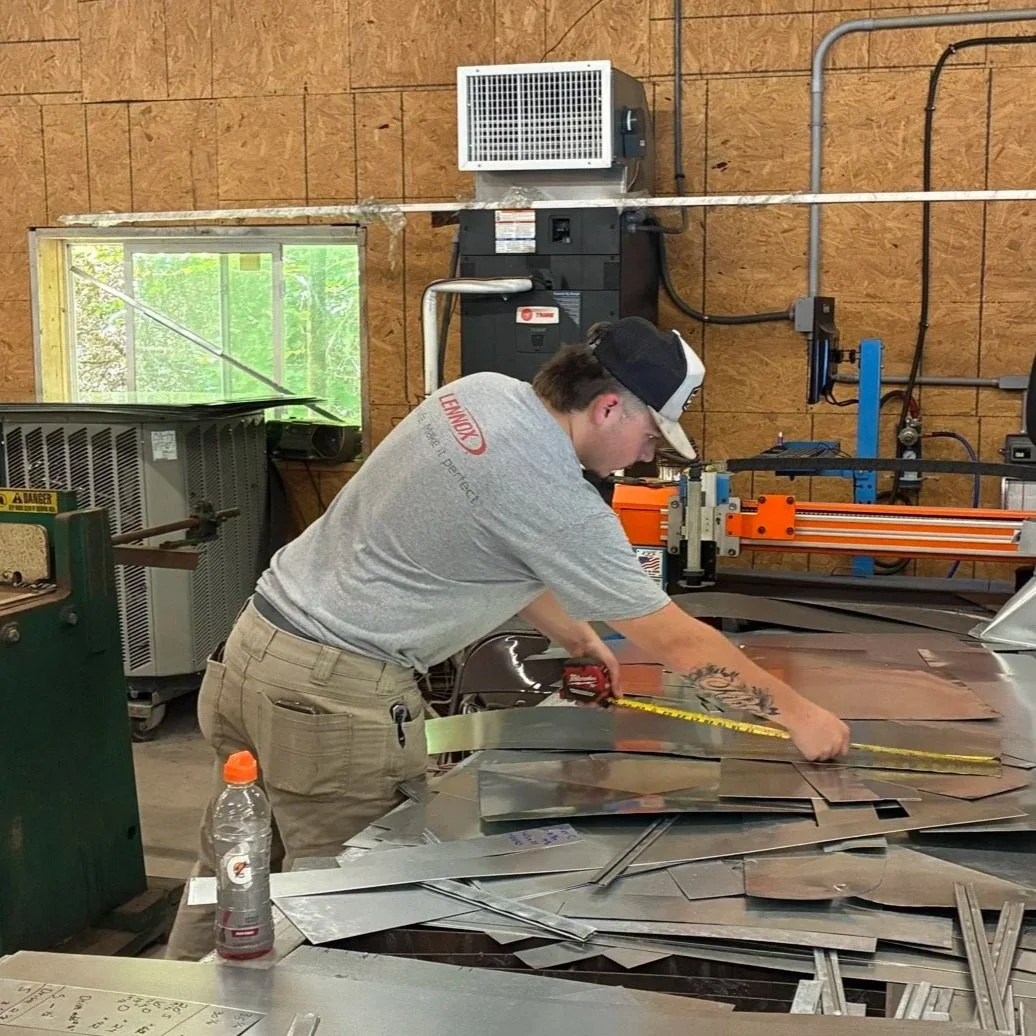 A man working in a metal workshop, measuring and cutting metal sheets on a workbench with a ruler. There are tools and metal pieces.