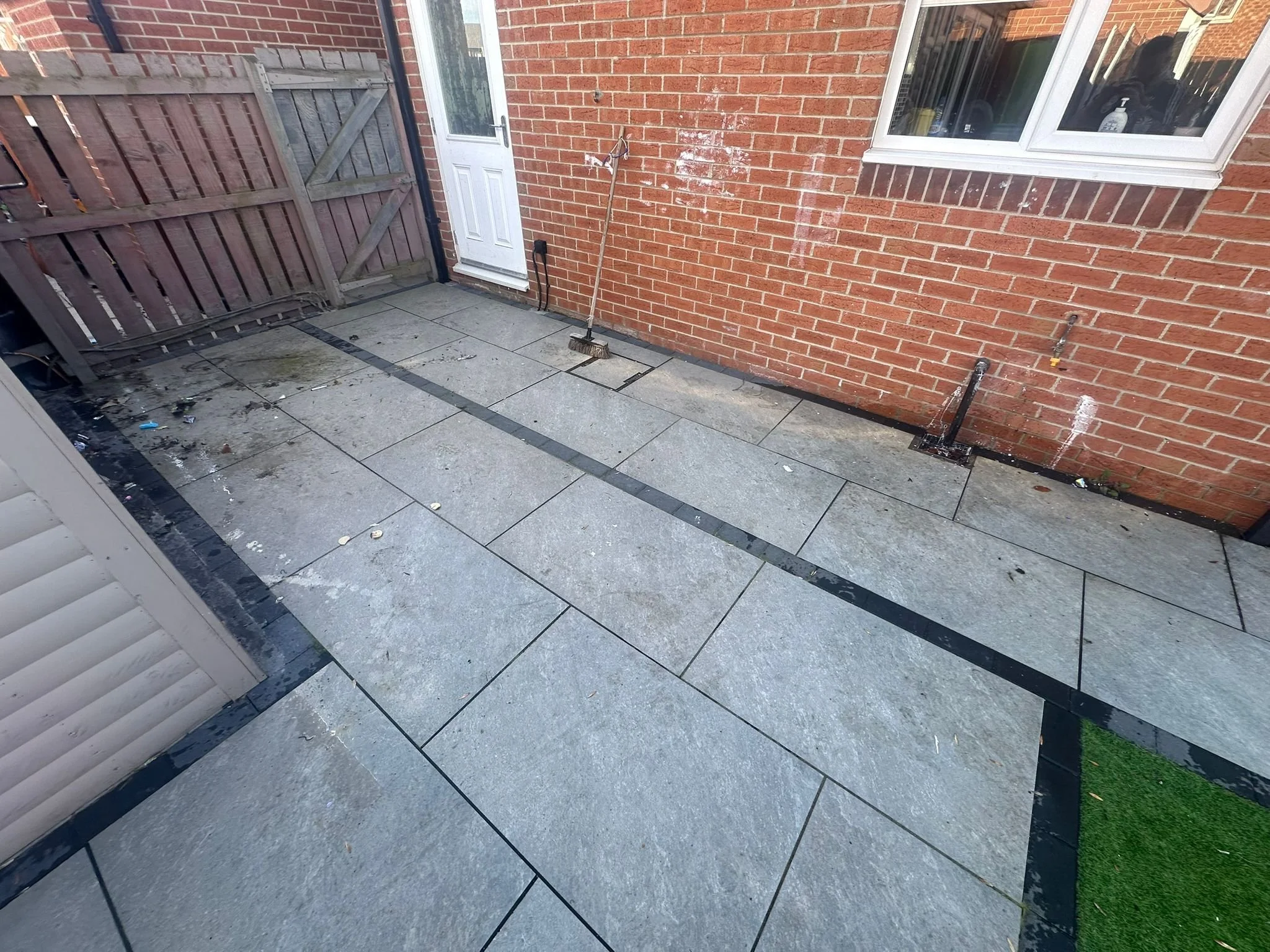 Backyard patio with tiled flooring, wooden fence, brick house wall, and a white door. There is a broom leaning against the wall and some scattered debris on the ground.