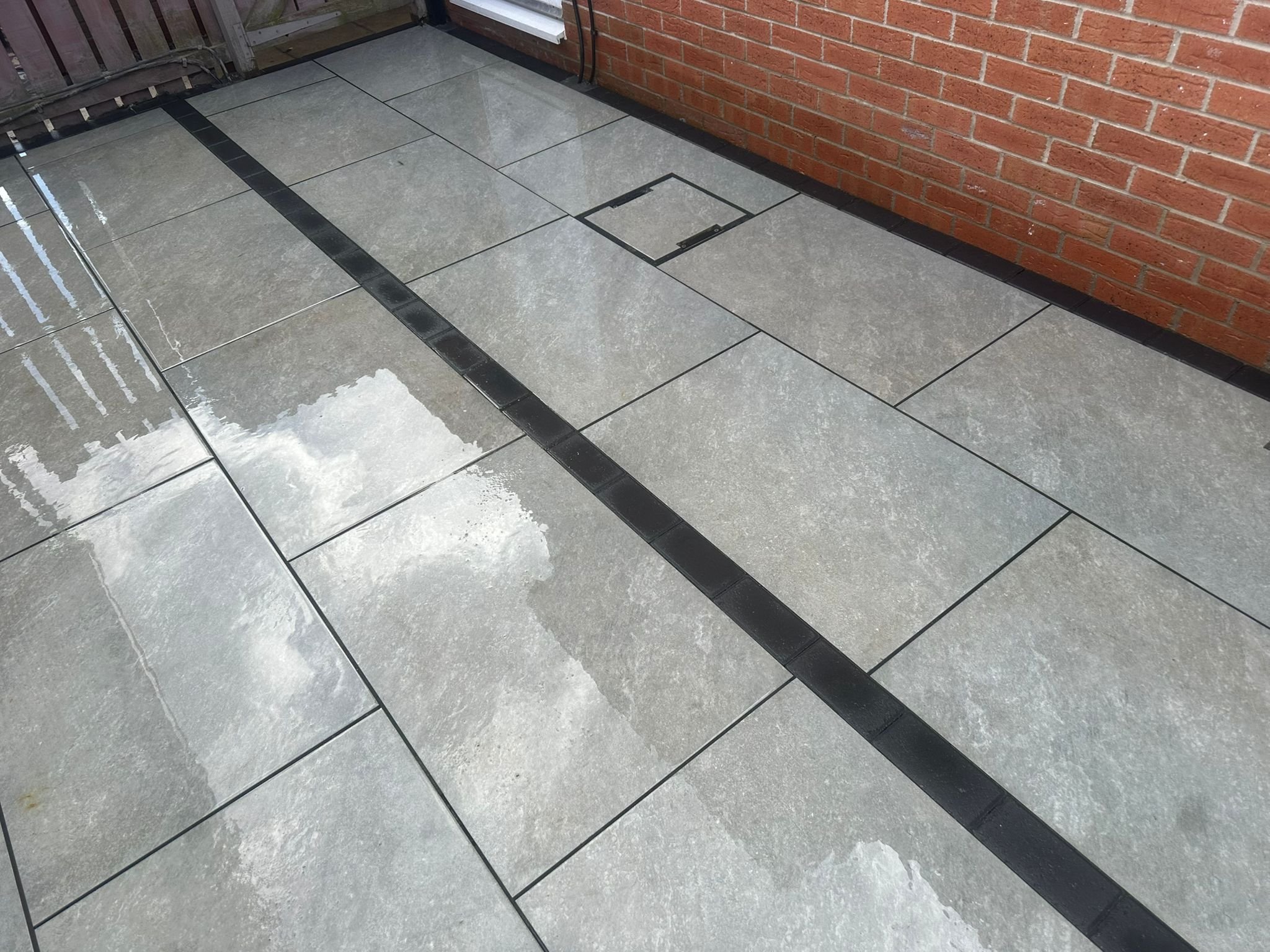 Wet gray tiled patio area bordered with black tiles, next to a red brick wall and a wooden fence.