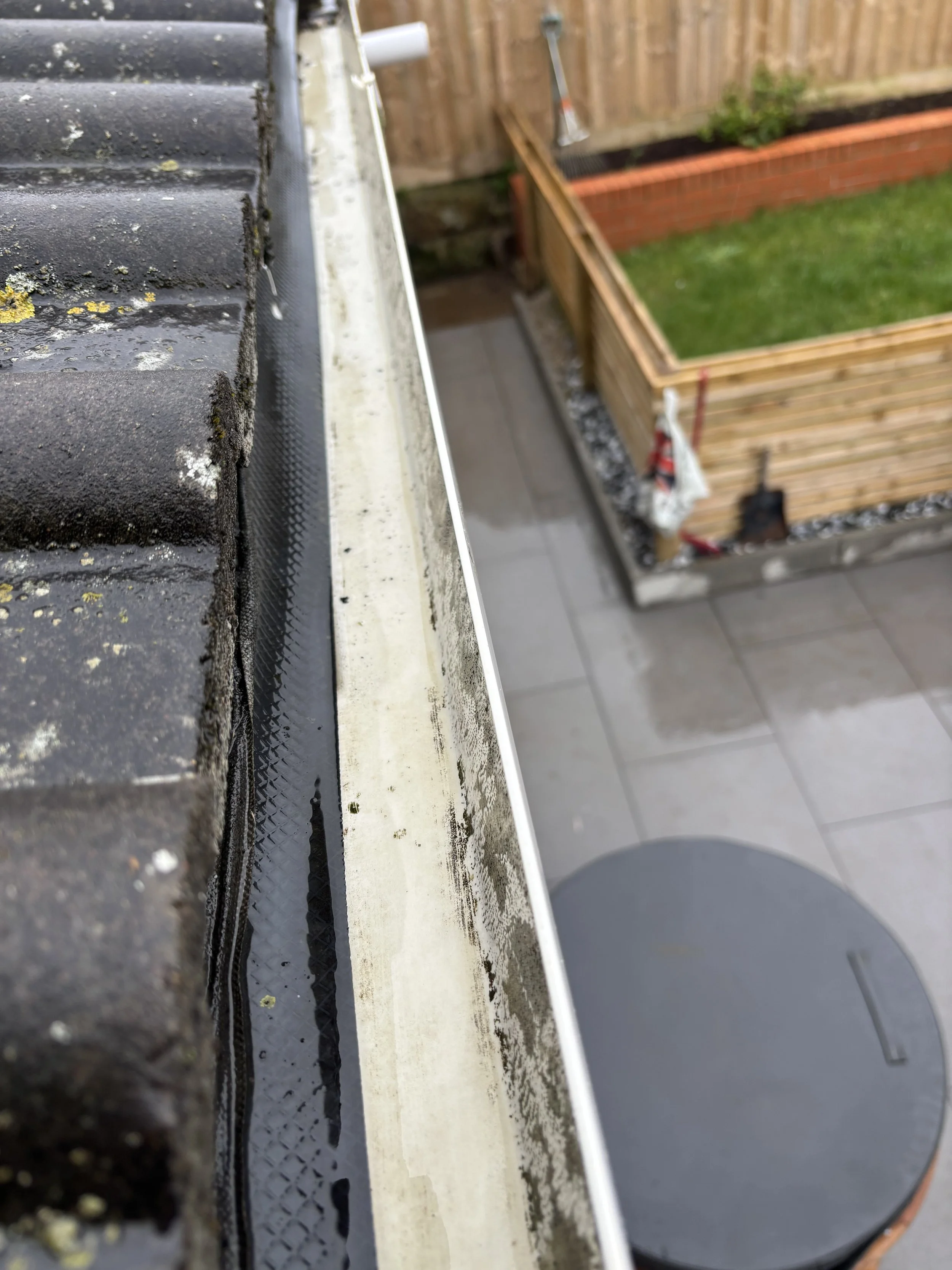 Close-up of a gutter with dirt, moss, and debris, with a blurred backyard garden with a wooden fence, raised planter bed, and gardening tools visible in the background.