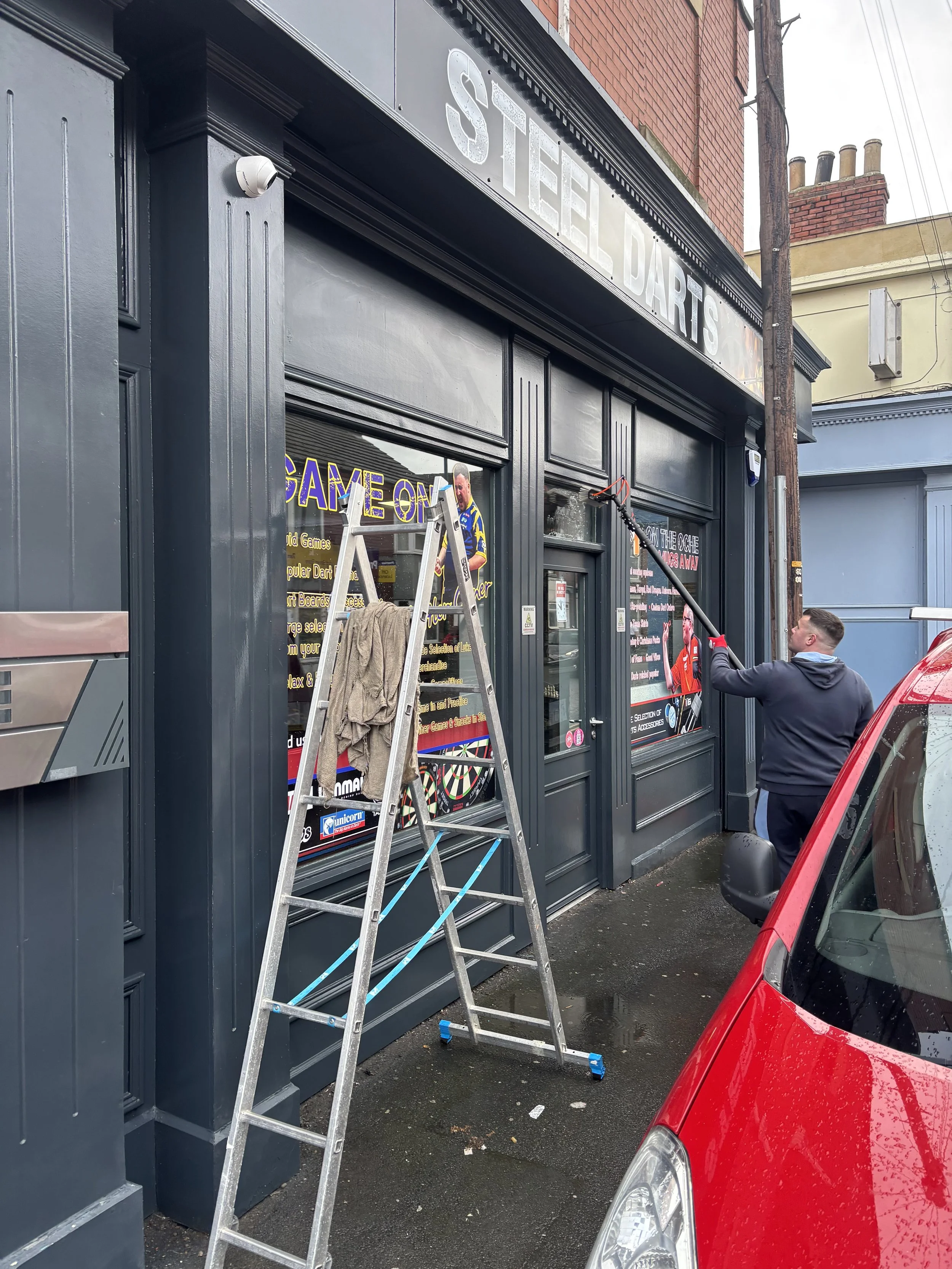 A person cleaning the exterior window of a dart shop with a spray and cloth, using a long-handled squeegee. The shop has black painted woodwork, and a ladder is leaning against the doorframe. A red car is parked in front of the shop.