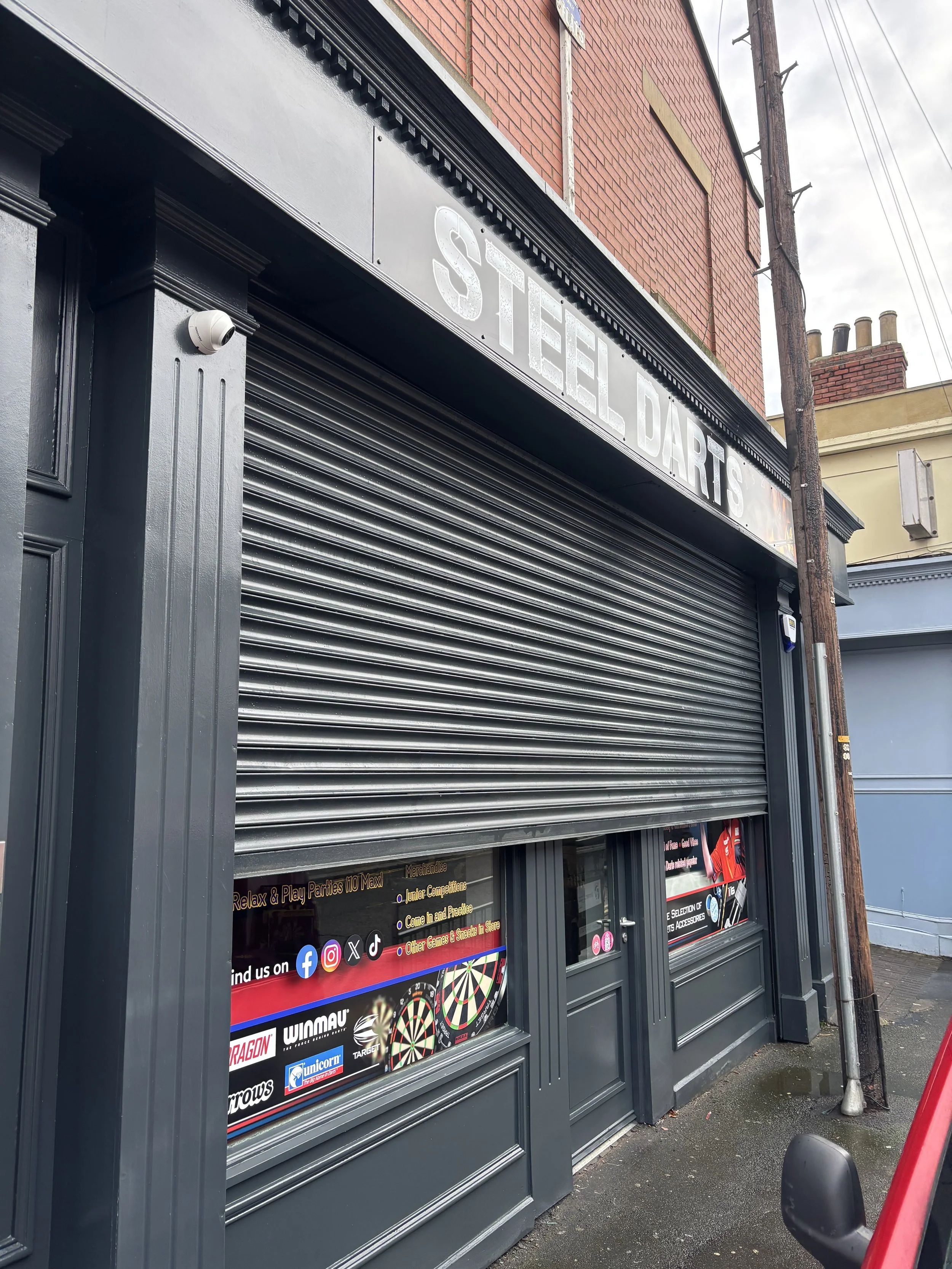 storefront with black and grey paint, partially closed metal shutter, and a window displaying advertisements for darts and games, outside a red brick building, next to a wooden utility pole and a parked scooter.