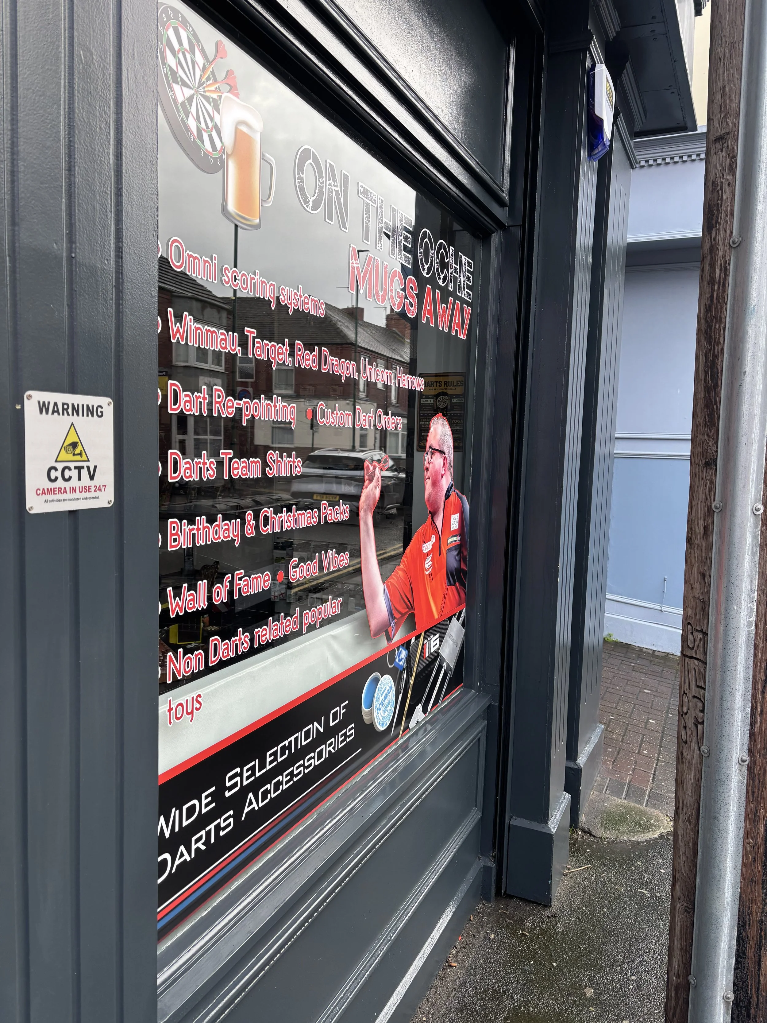 A storefront window advertising a dart shop, with a dartboard graphic and a man in a red shirt holding a dart, signage listing products like dart scoring systems, darts, and accessories, and a CCTV warning sign on the side.