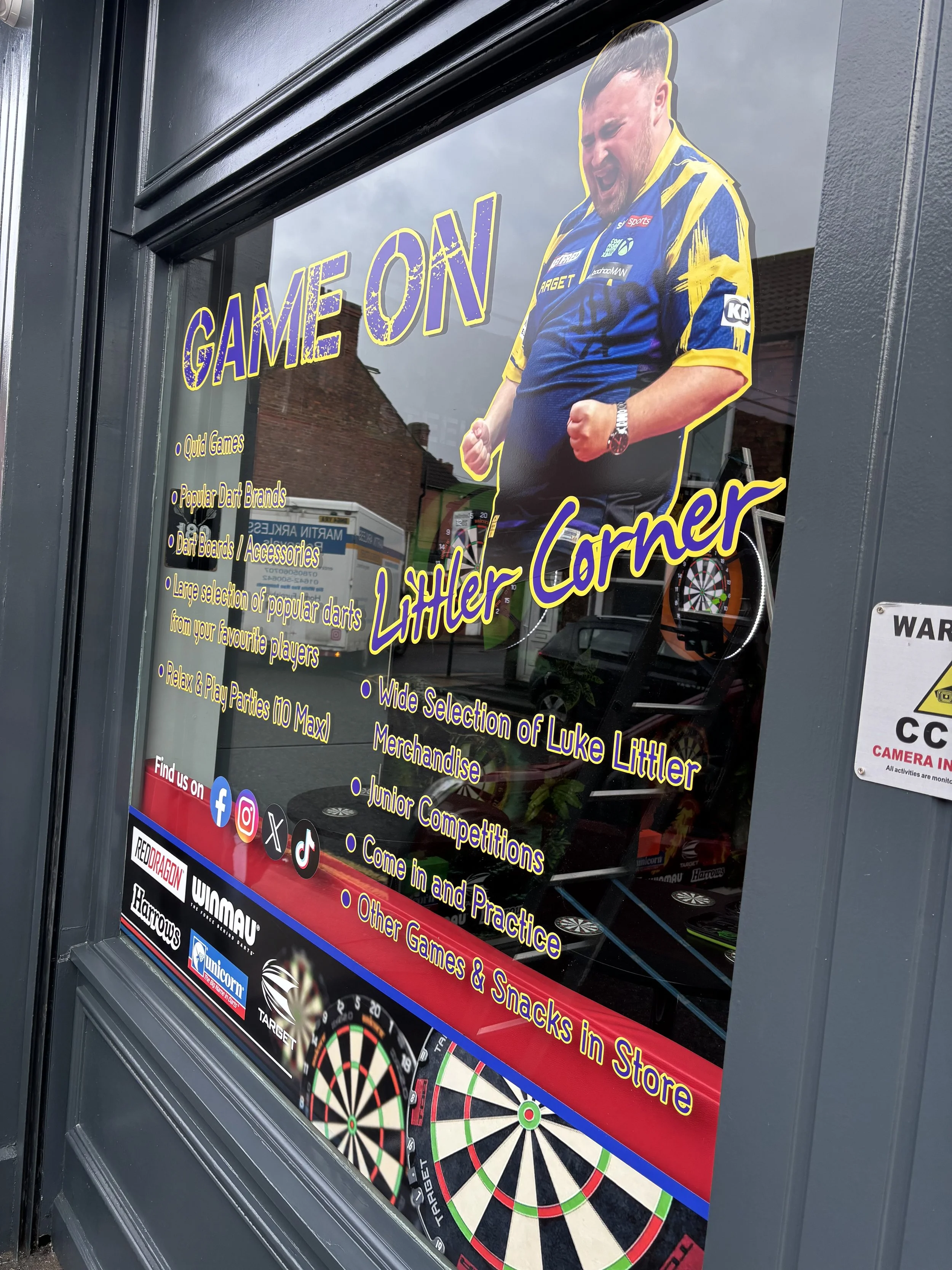 A shop window with a poster advertising dart games and equipment, featuring a man in a blue and yellow sports jersey celebrating with fists clenched, standing in front of dartboards.