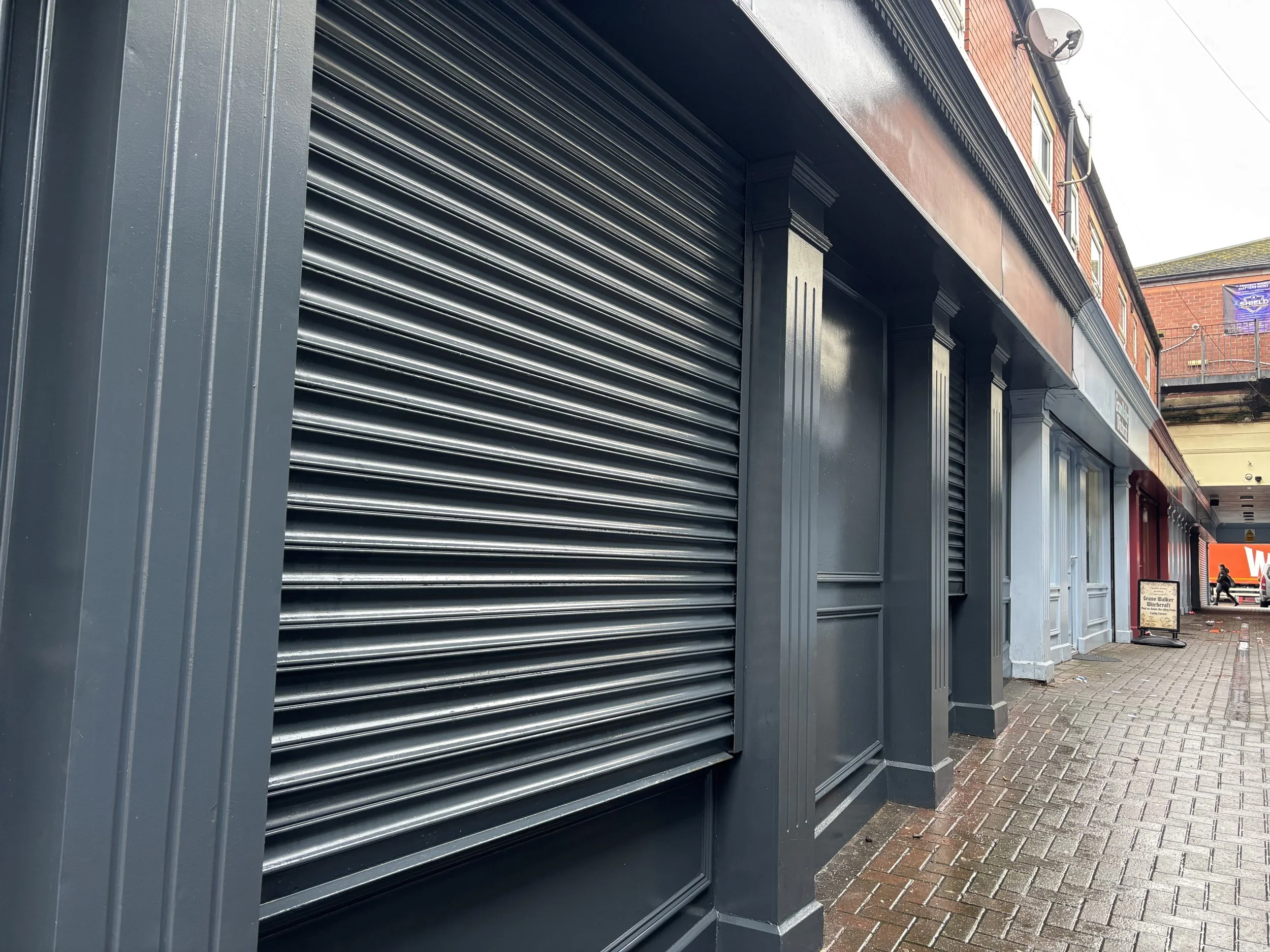 Closed storefront with a black metallic roll-up shutter, decorative gray columns, and red brick building above. Street scene with a sidewalk and a signboard in the background.