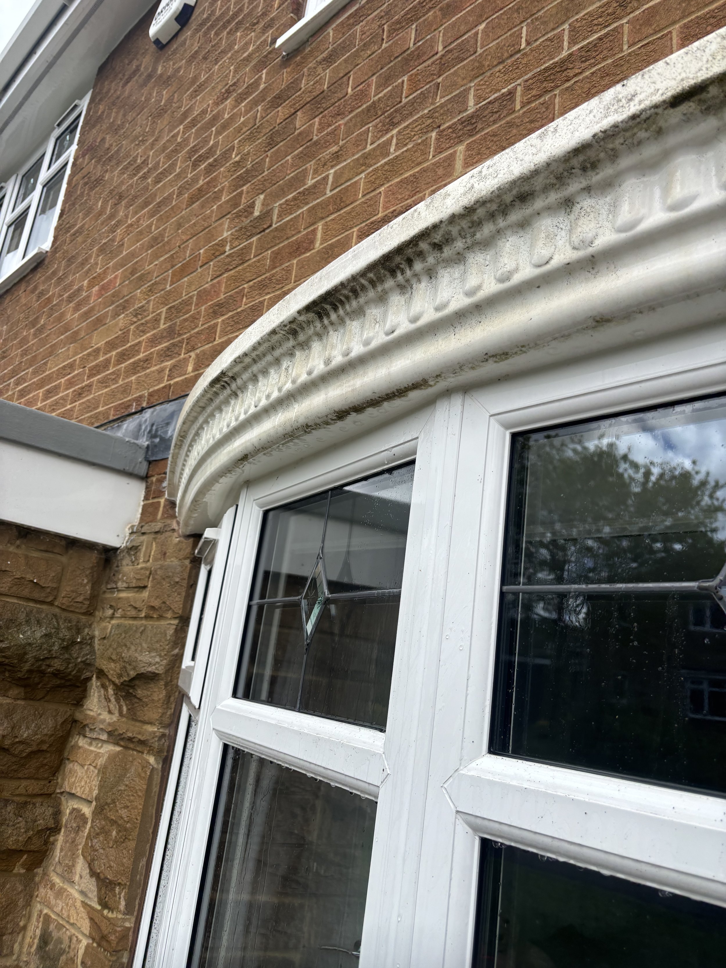 Close-up photo of a white double-glazed window with a decorative diamond pattern on the inner glass panel, set into a red brick wall with some moss or dirt on the window frame.