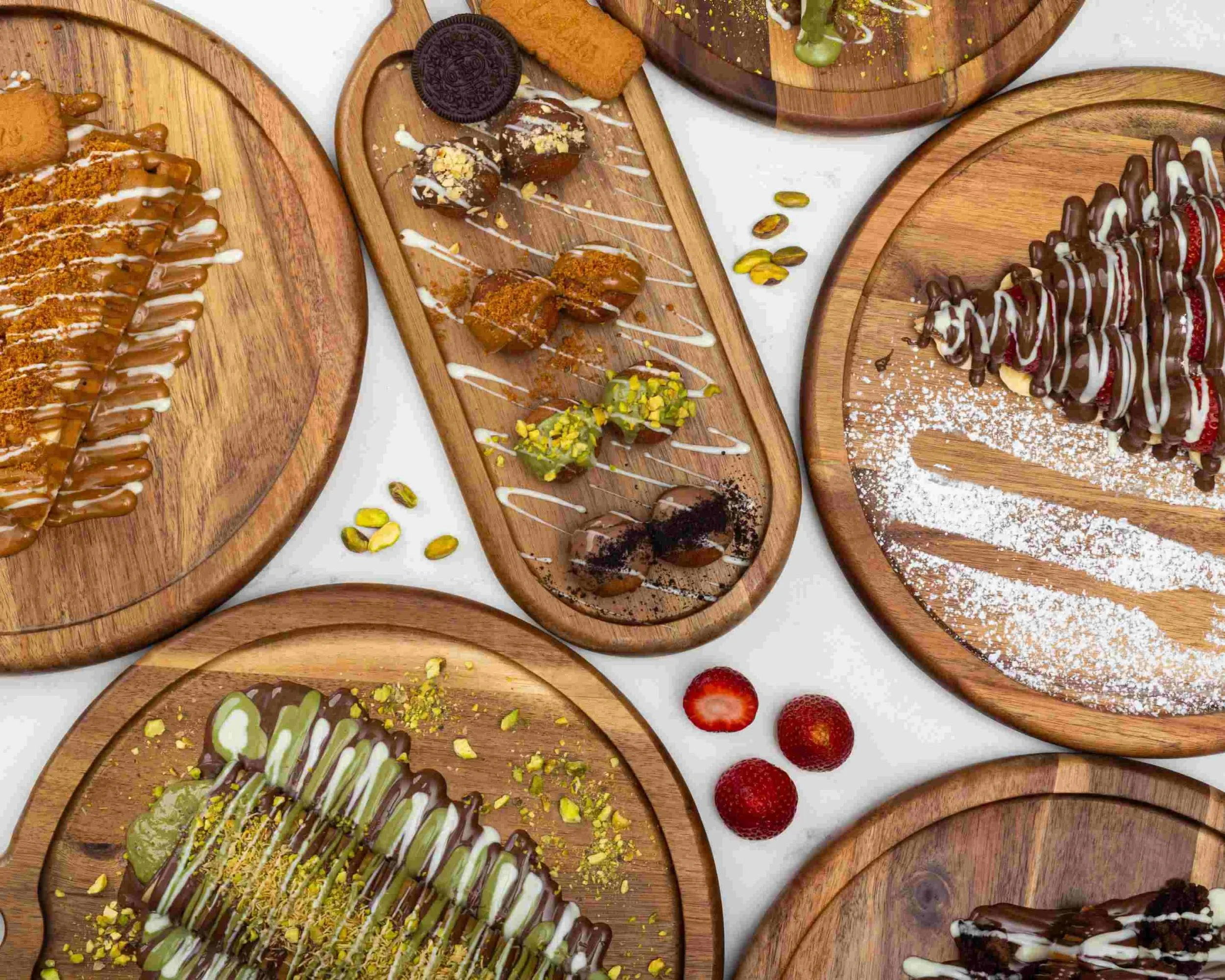 Assorted desserts including a chocolate-covered pastry, assorted chocolate truffles, a banana split with chocolate and white drizzle, and strawberries on wooden plates.