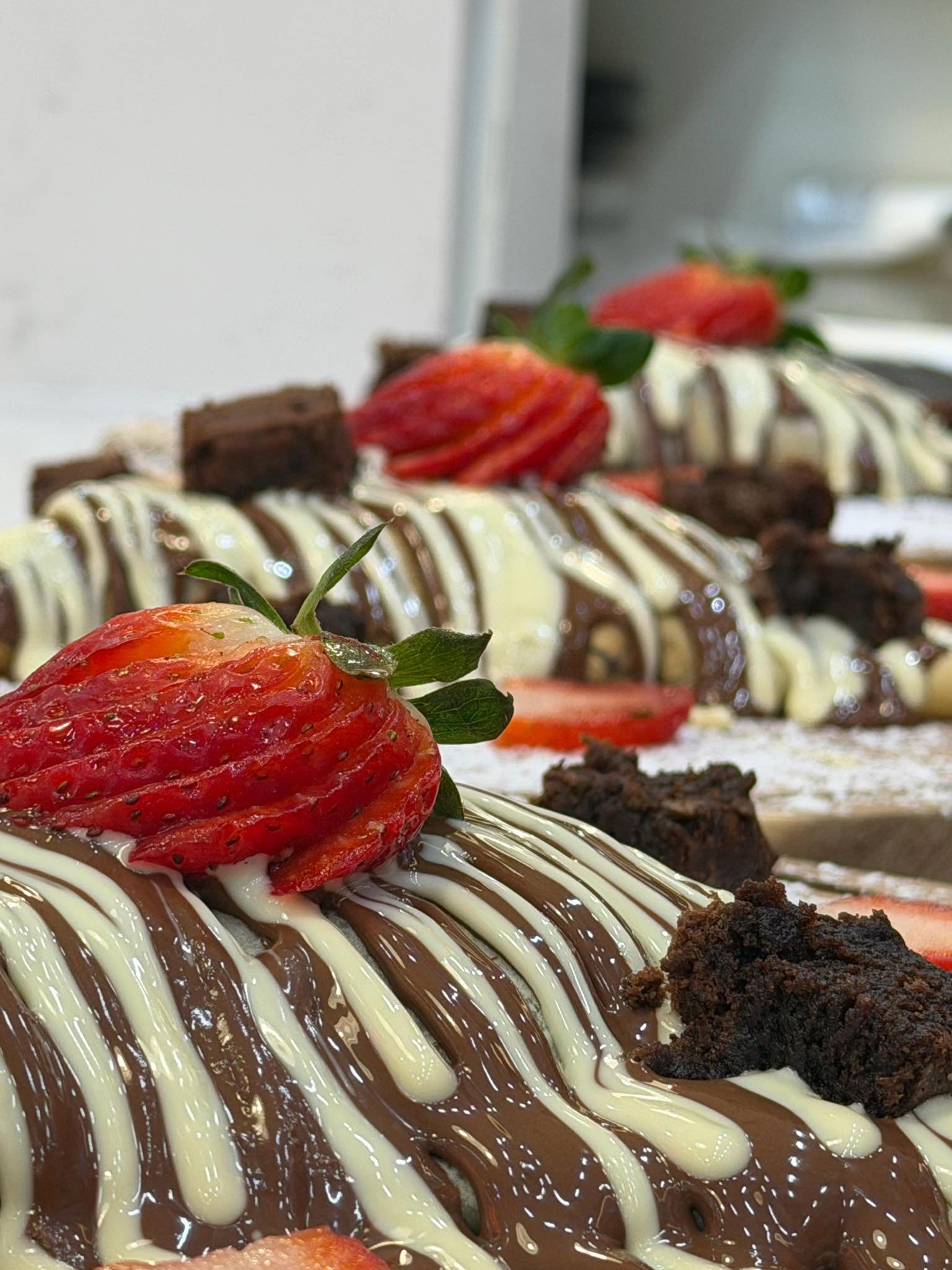 Close-up of decorated chocolate cake slices topped with sliced strawberries, white and dark chocolate drizzle, and small brownie pieces on a plate.
