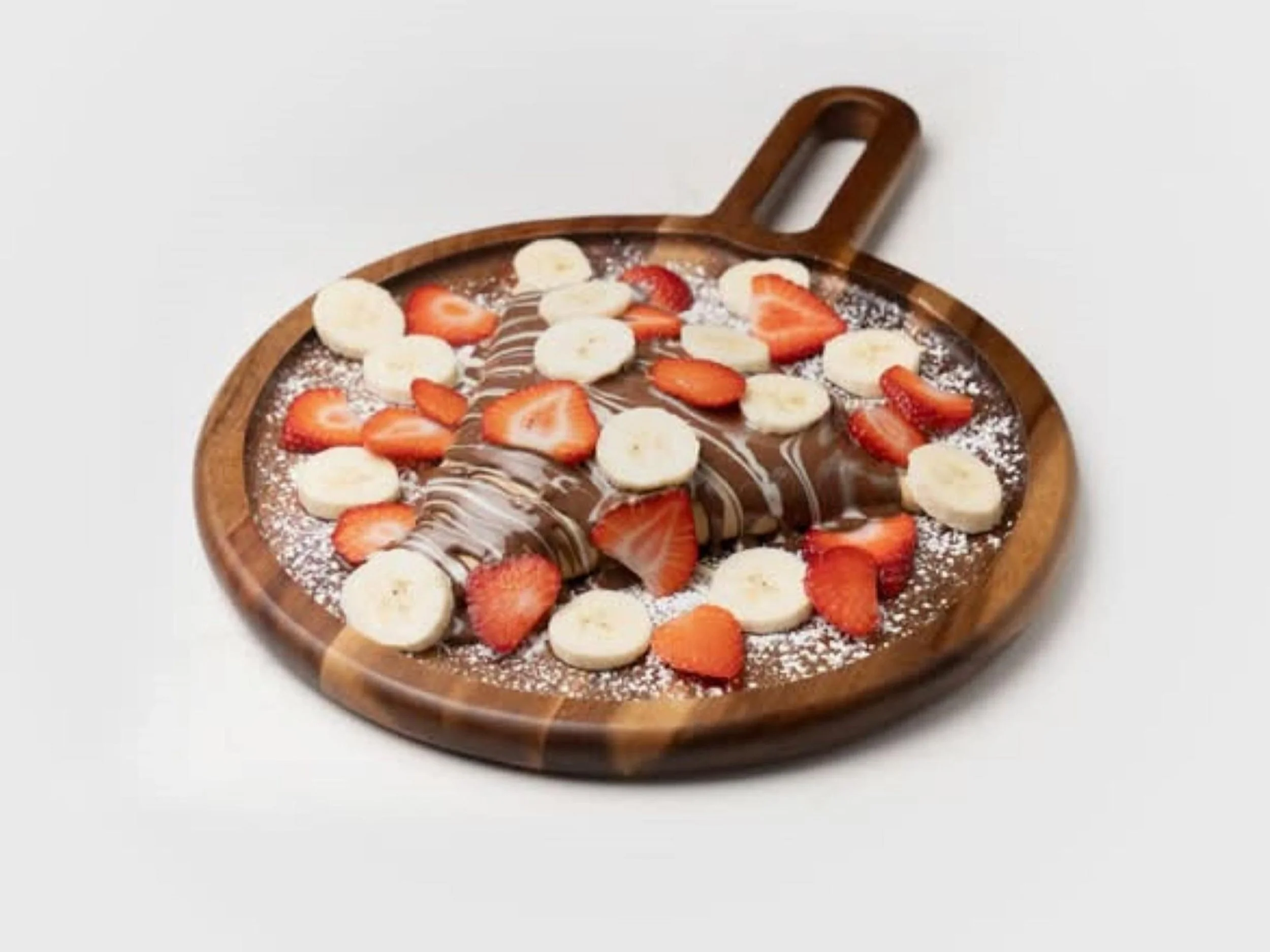 A wooden serving board with a chocolate crepe topped with banana slices, strawberry slices, and white powdered sugar.