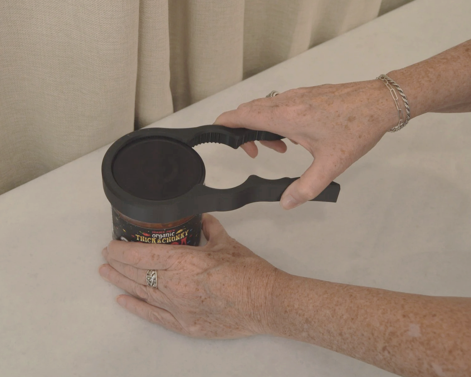 A person with freckled hands and silver jewelry uses a can opener to open a jar of organic thick and chunky tomato sauce, placed on a white surface near beige curtains.