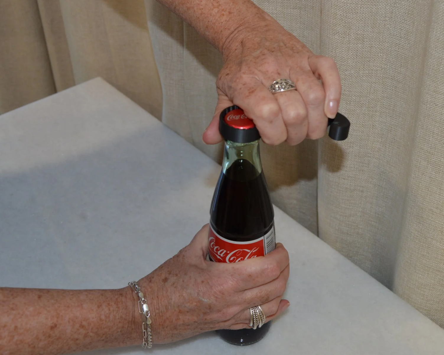 Hand opening a mini Coca-Cola glass bottle with a metal cap using a bottle opener.