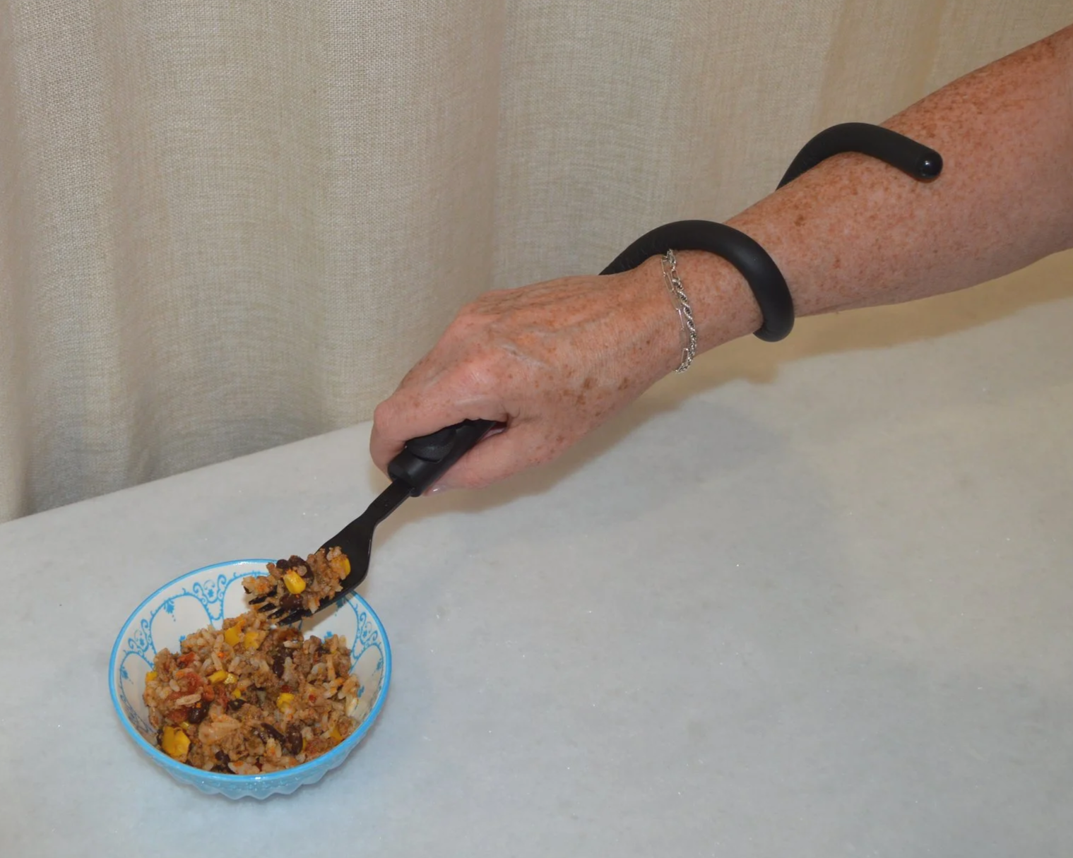 A hand with a black bracelet, a silver bracelet, and freckled skin, holding a black fork, serving a portion of taco salad from a small blue bowl on a white surface.