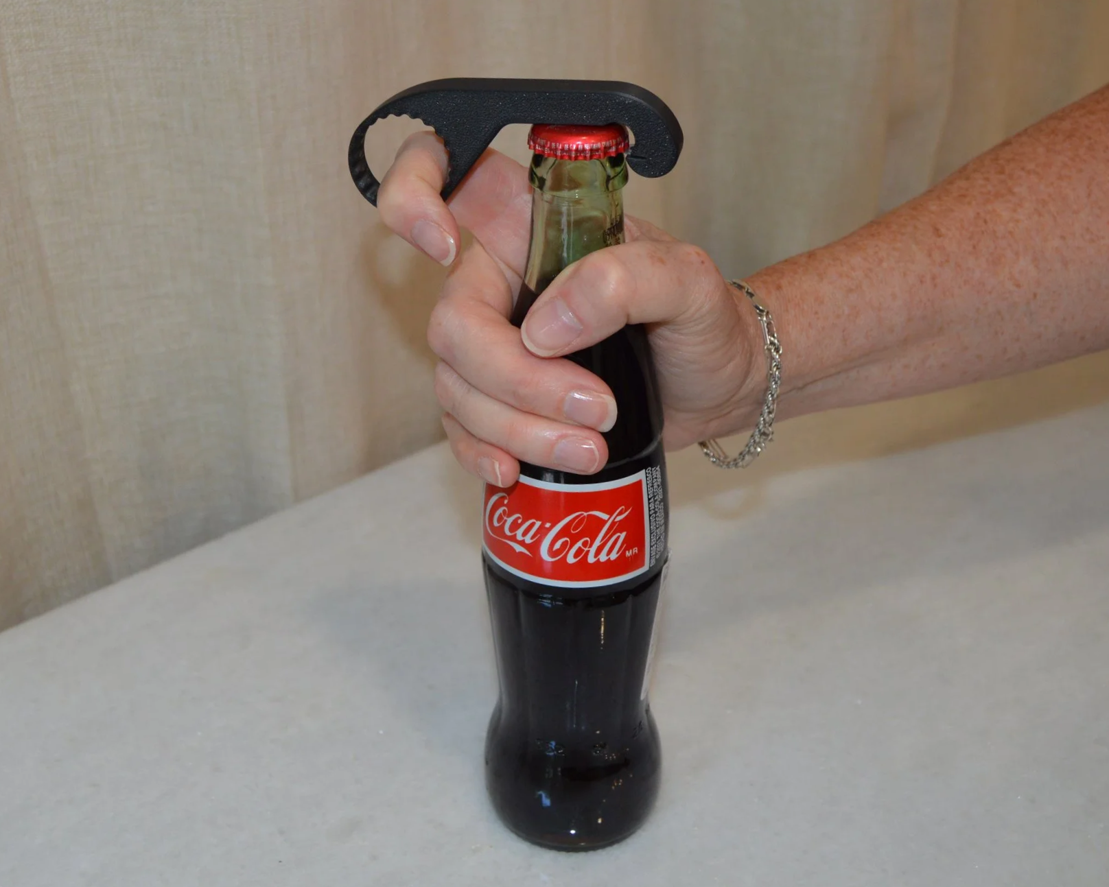 Hand using a bottle opener on a Coca-Cola glass bottle on a beige counter.