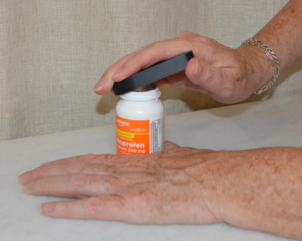 A person opening a bottle of ibuprofen tablets with their right hand, while resting their left hand on a flat surface.