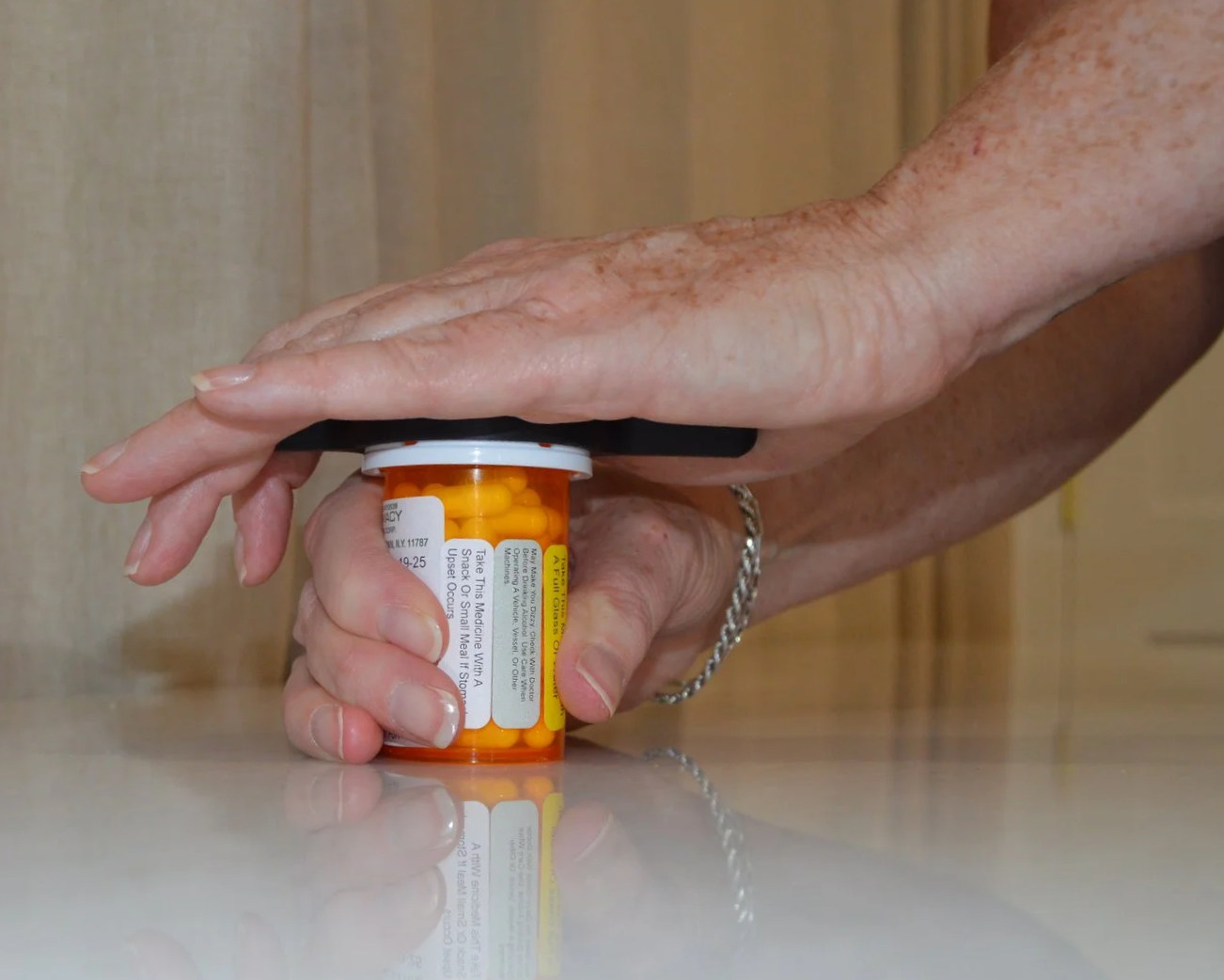 A person's hand pressing a pill bottle with yellow capsules on a white marble countertop.