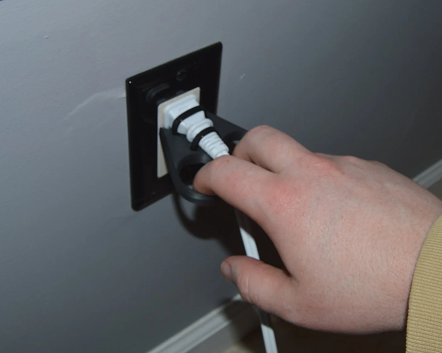 A hand unplugging a white electrical cord from a wall outlet on a gray wall.
