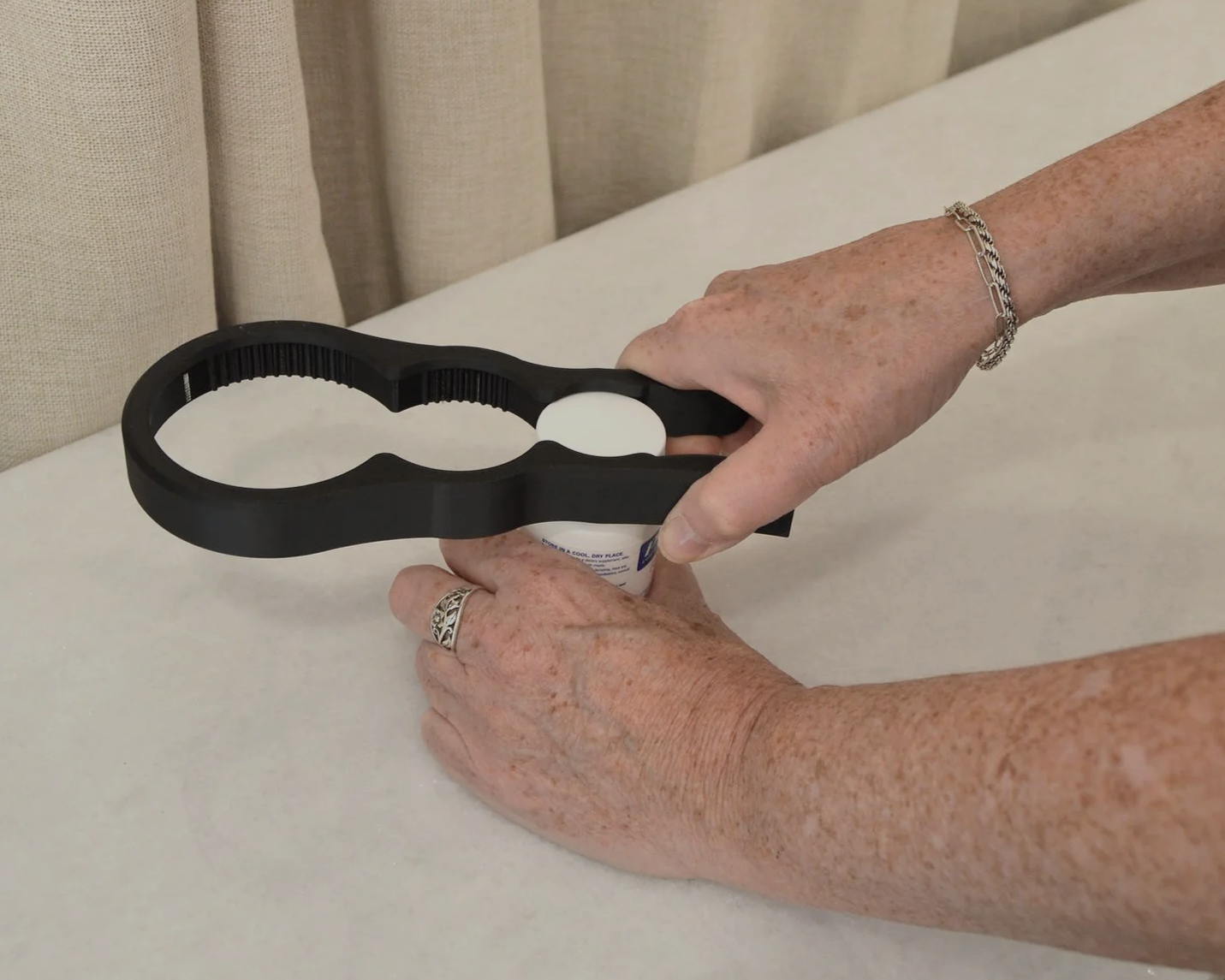 A person with freckled arms wearing rings and a bracelet is placing a black, hand-held device over a container on a white surface.