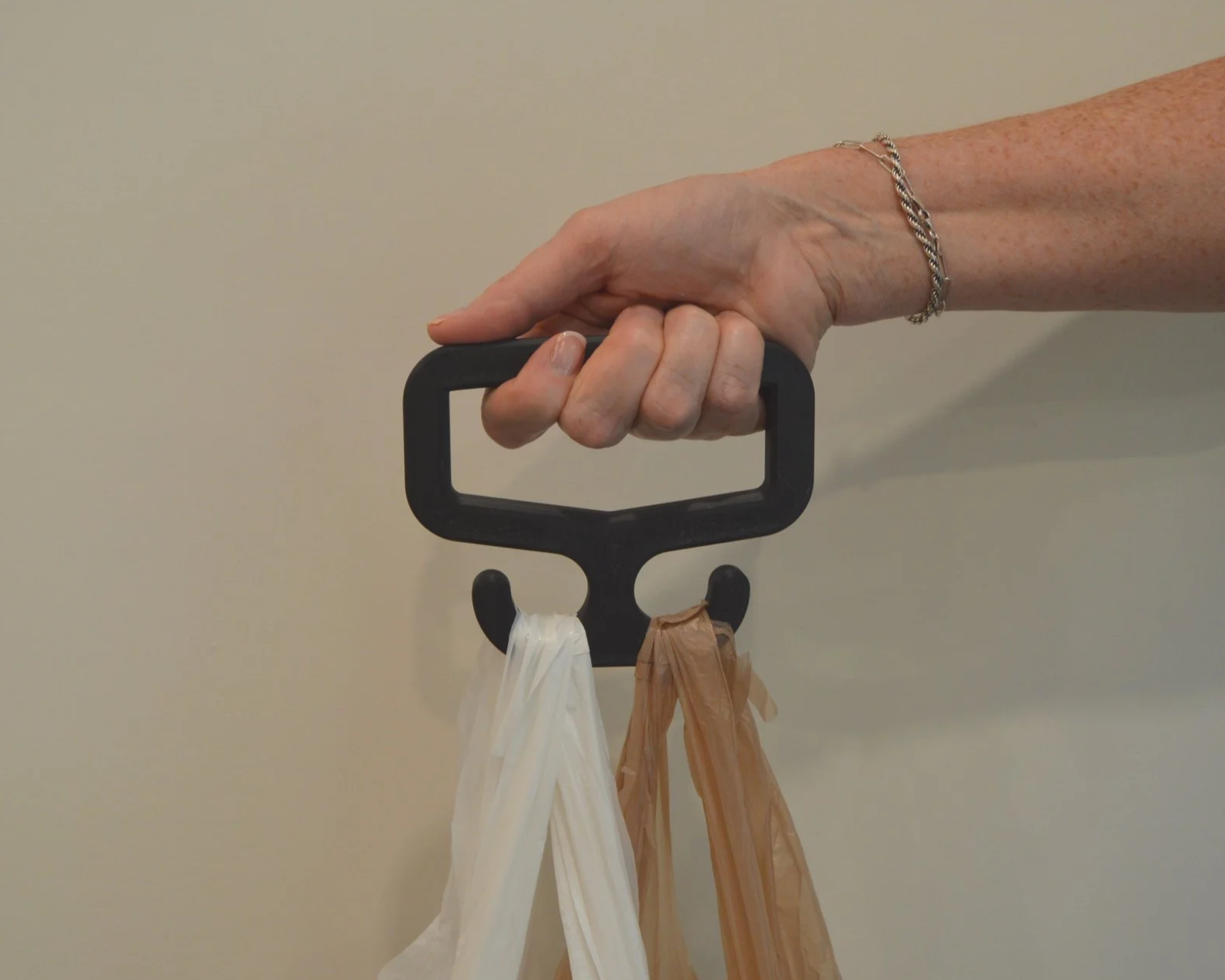 A hand holding a black wall hook with plastic and paper bags hanging from it, against a plain beige wall.