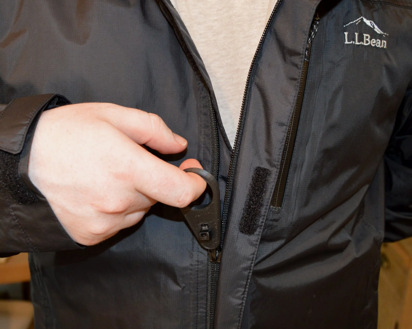 A person wearing a gray L.L.Bean jacket is using a black bottle opener to open a black zipper on the jacket.
