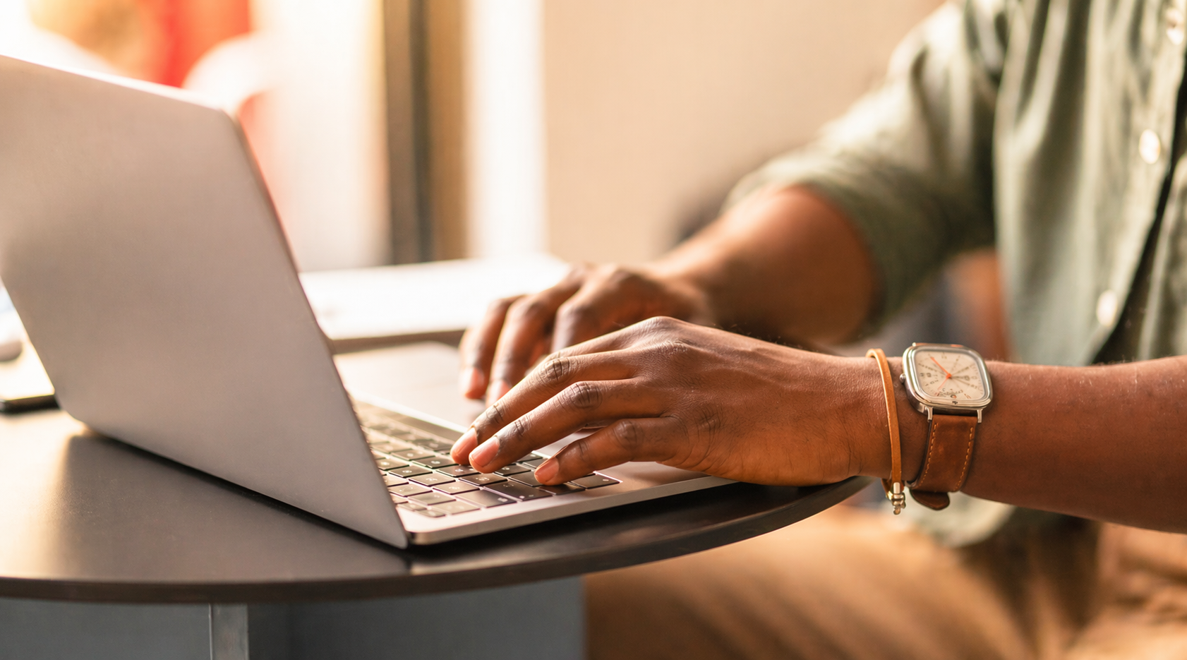 A person typing on a silver laptop while sitting at a black table. The person is wearing a wristwatch with a leather strap.