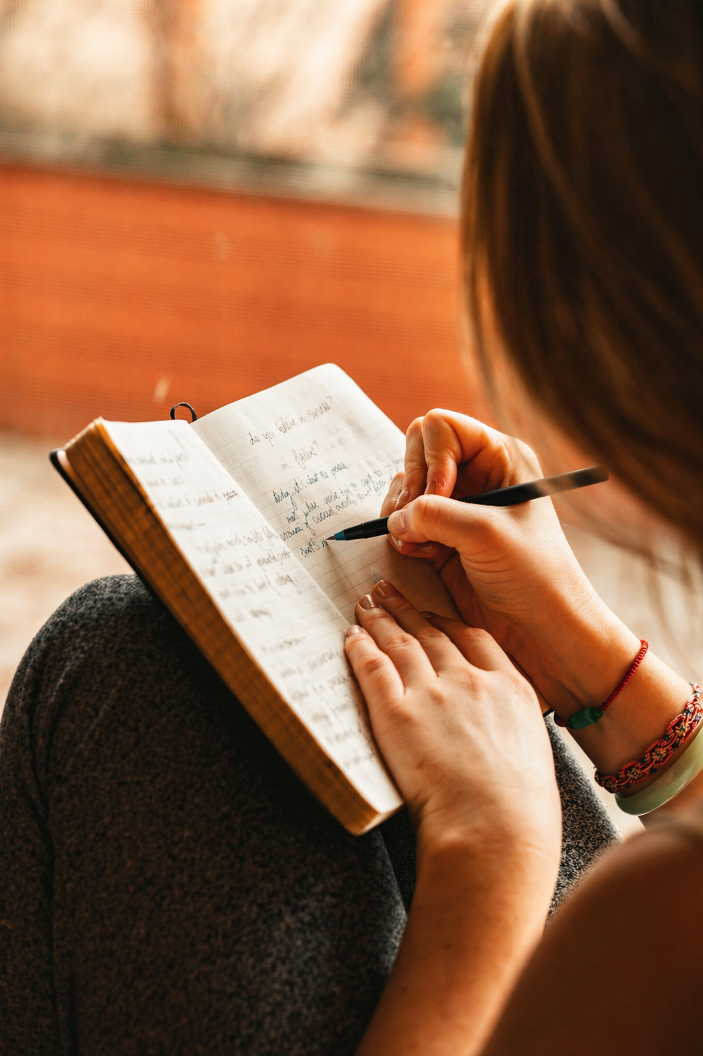 A person sitting outdoors, writing in a notebook with a pen, wearing multiple bracelets, with a red brick wall in the background.