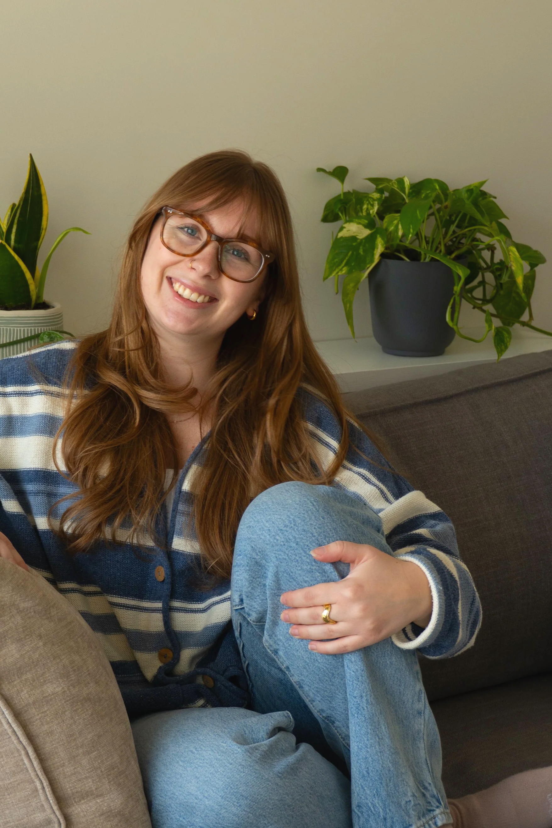 A young woman with long red hair, glasses, and a nose piercing sitting on a sofa, smiling, wearing a striped jacket and blue jeans, with green houseplants in the background.