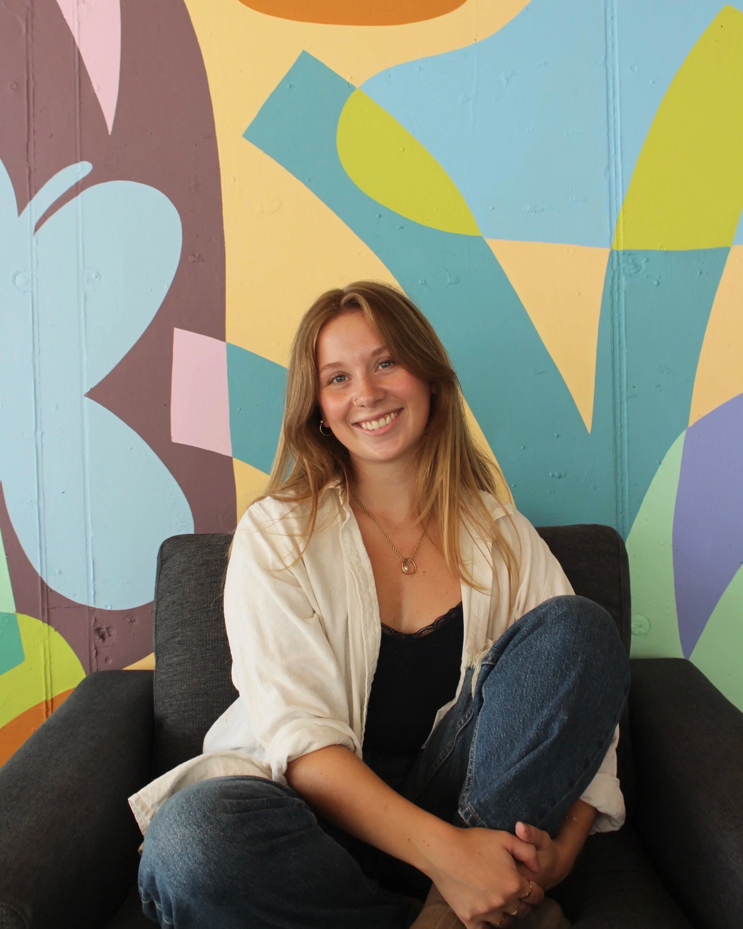 A young woman with long blonde hair, smiling, sitting in front of a colorful, abstract mural on a wall, wearing a white shirt and blue jeans.