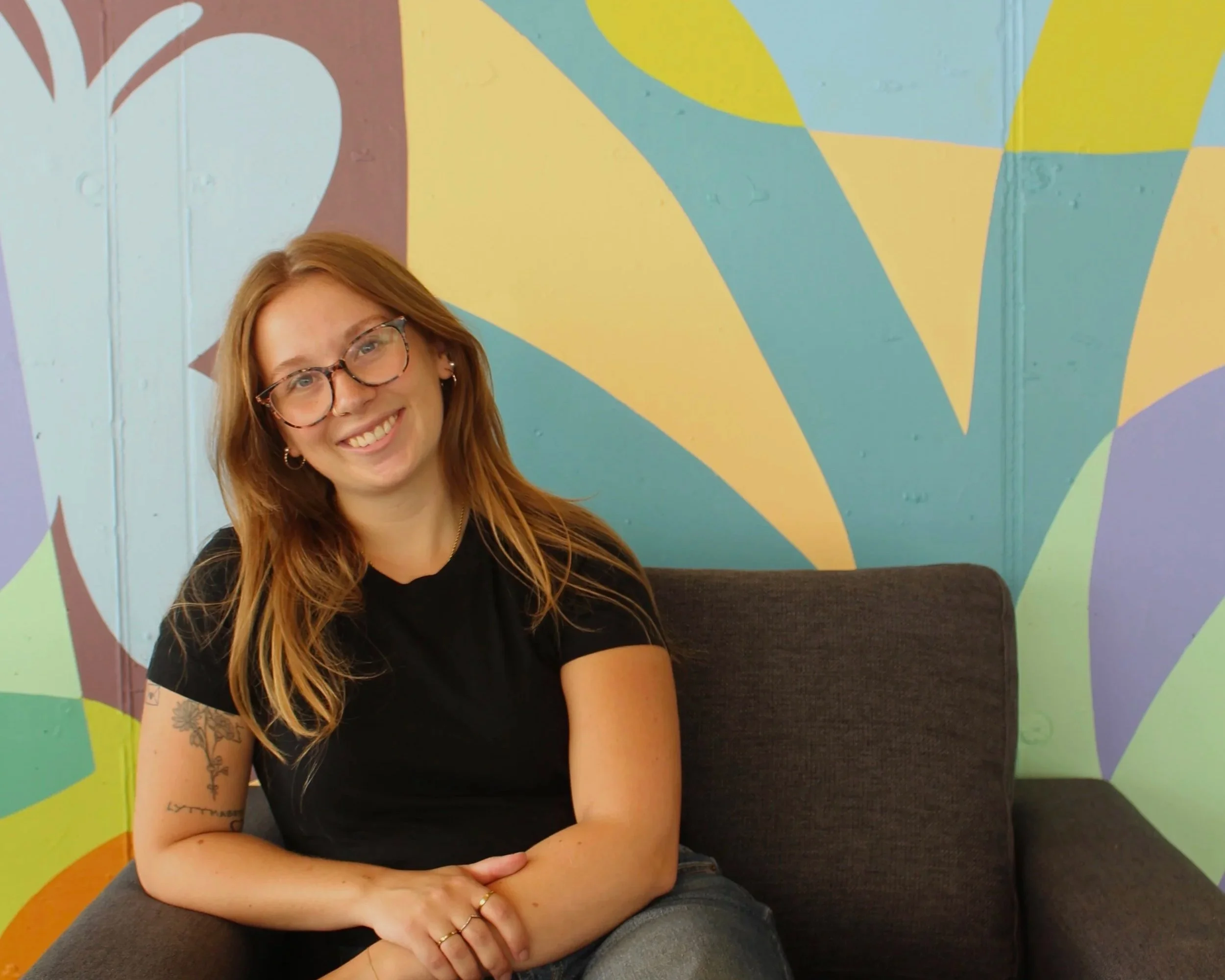 Blonde woman smiling, sitting in front of multi-coloured background.