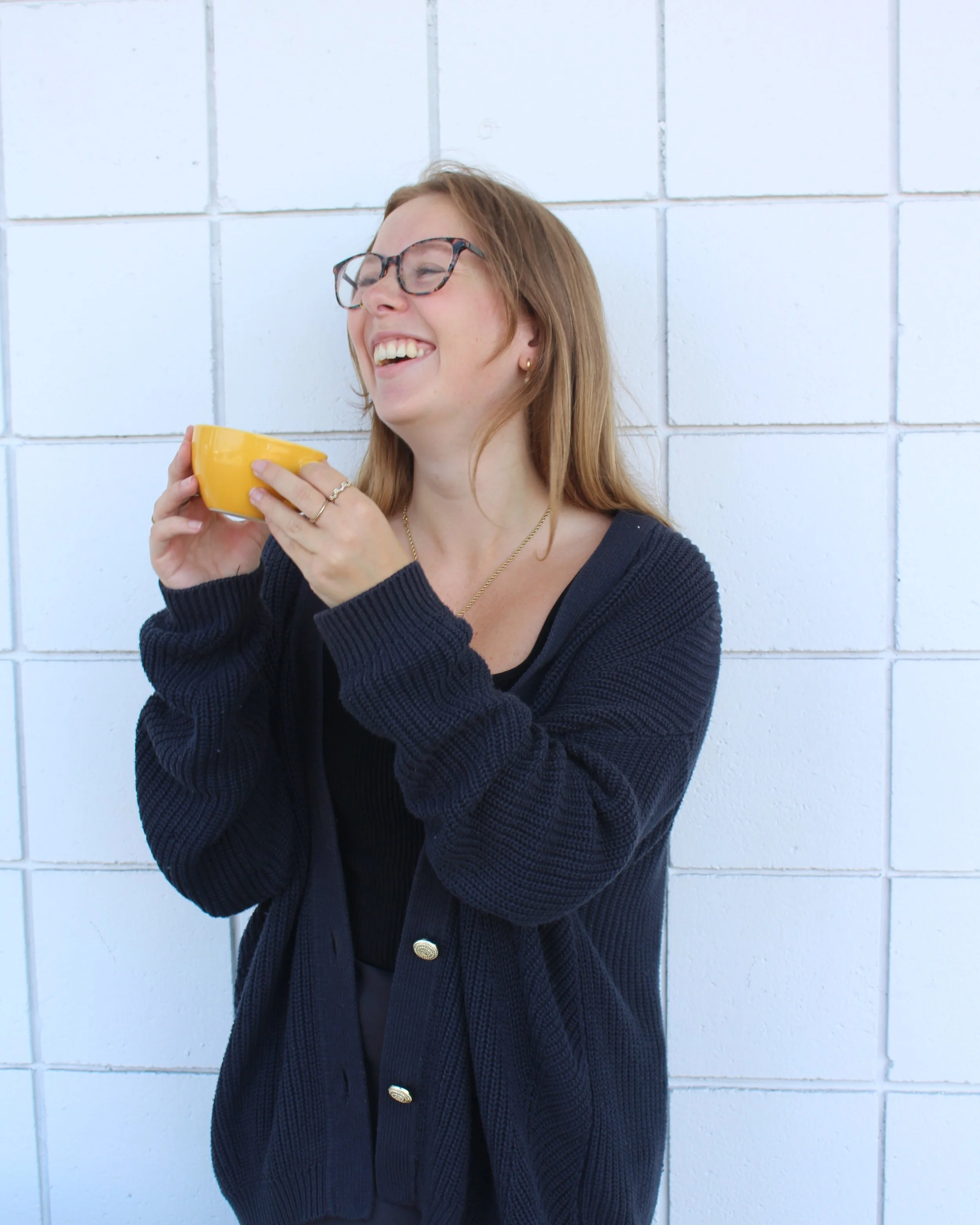 A woman with glasses and long red hair smiling and holding a yellow mug, standing in front of a white tiled wall.