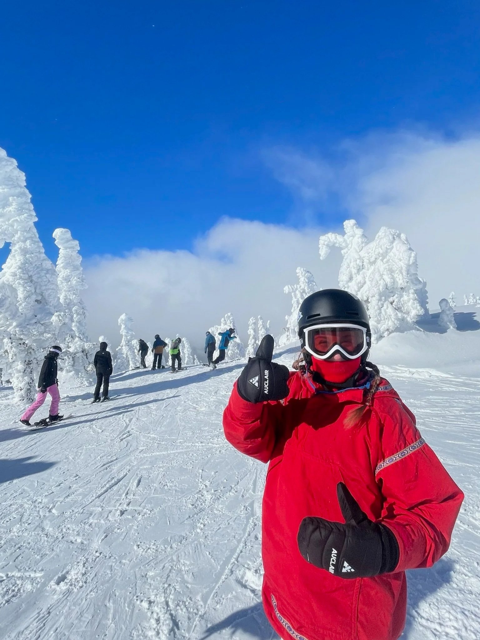 A person in red winter gear, black helmet, and ski goggles giving a thumbs up on a snowy mountain slope. Several other skiers and snowboarders are in the background, with snow-covered trees and a clear blue sky.